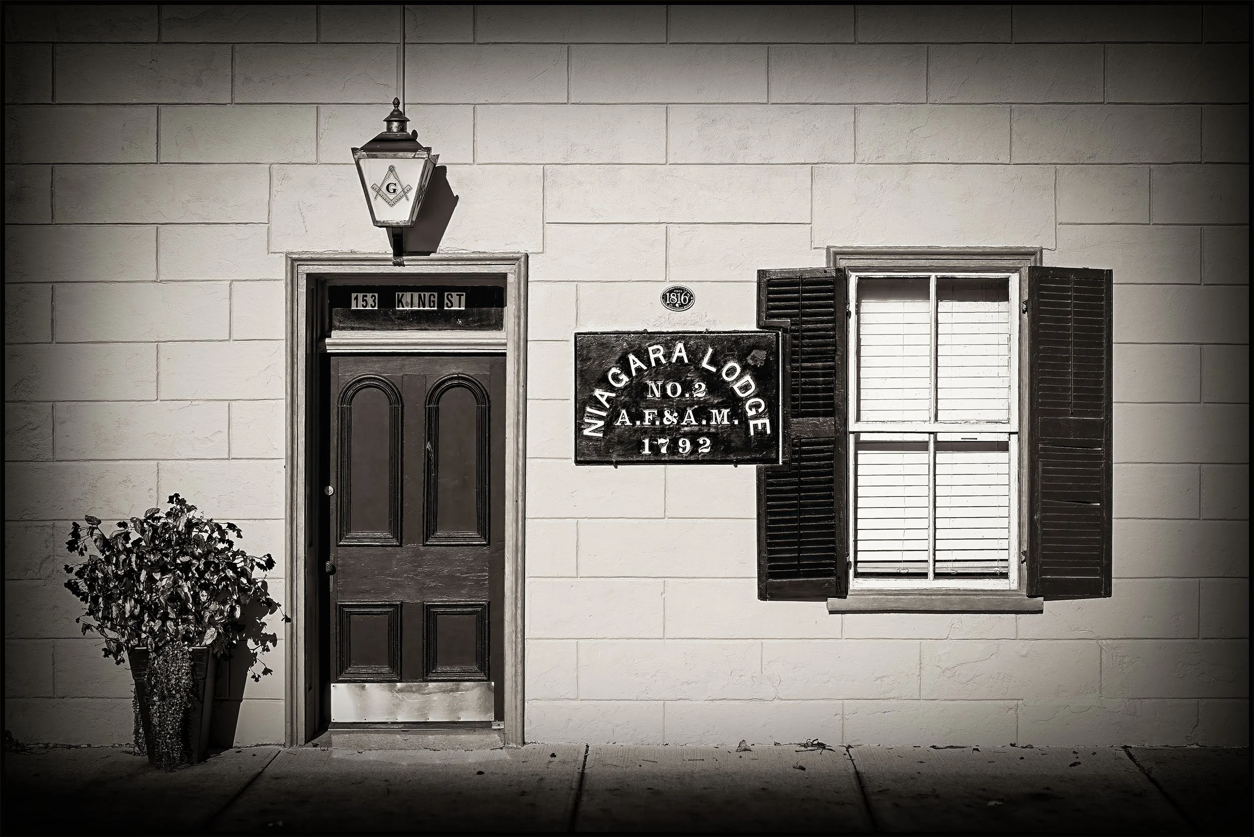 A black and white photo of a building's front wall with a closed door, a window with partly open shutters, a lamp above the door, and a sign indicating the address and historical details.