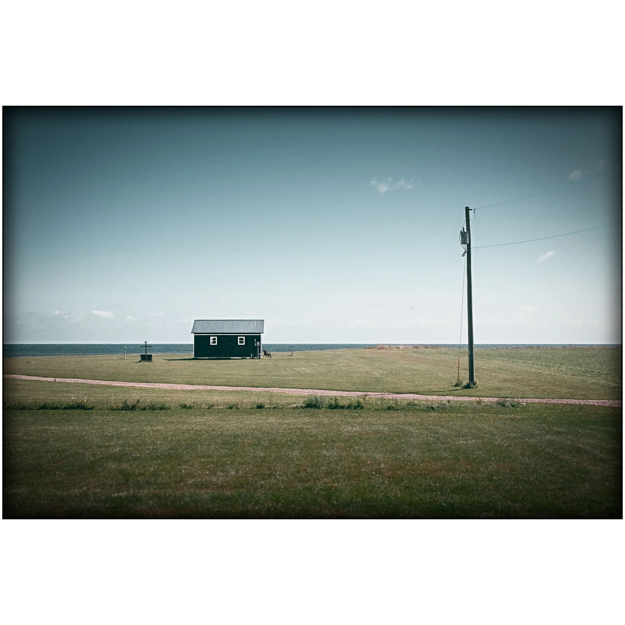 A solitary dark cabin sits in a wide grassy field near the shoreline, with a utility pole to one side and open water on the horizon.