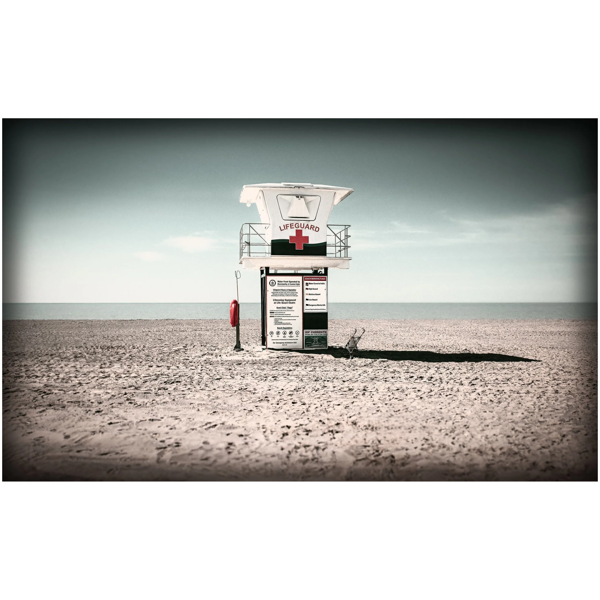 A white lifeguard stand with red cross markings sits alone on a sandy beach, with calm water and a pale horizon behind it.