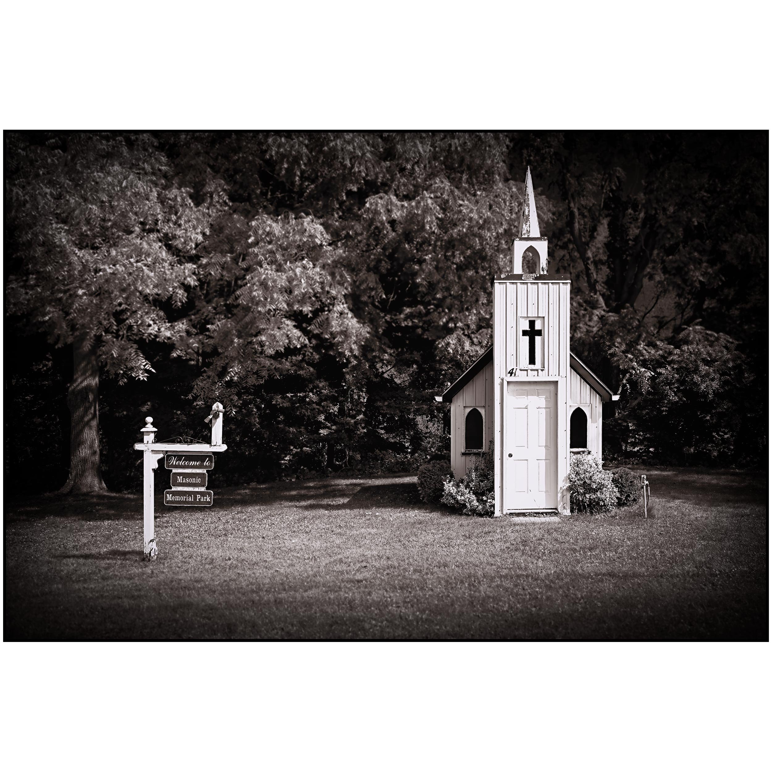 Black and white photograph of a small church with a steeple and cross, surrounded by trees, with a 'Welcome to Masonic Memorial Park' sign in front.