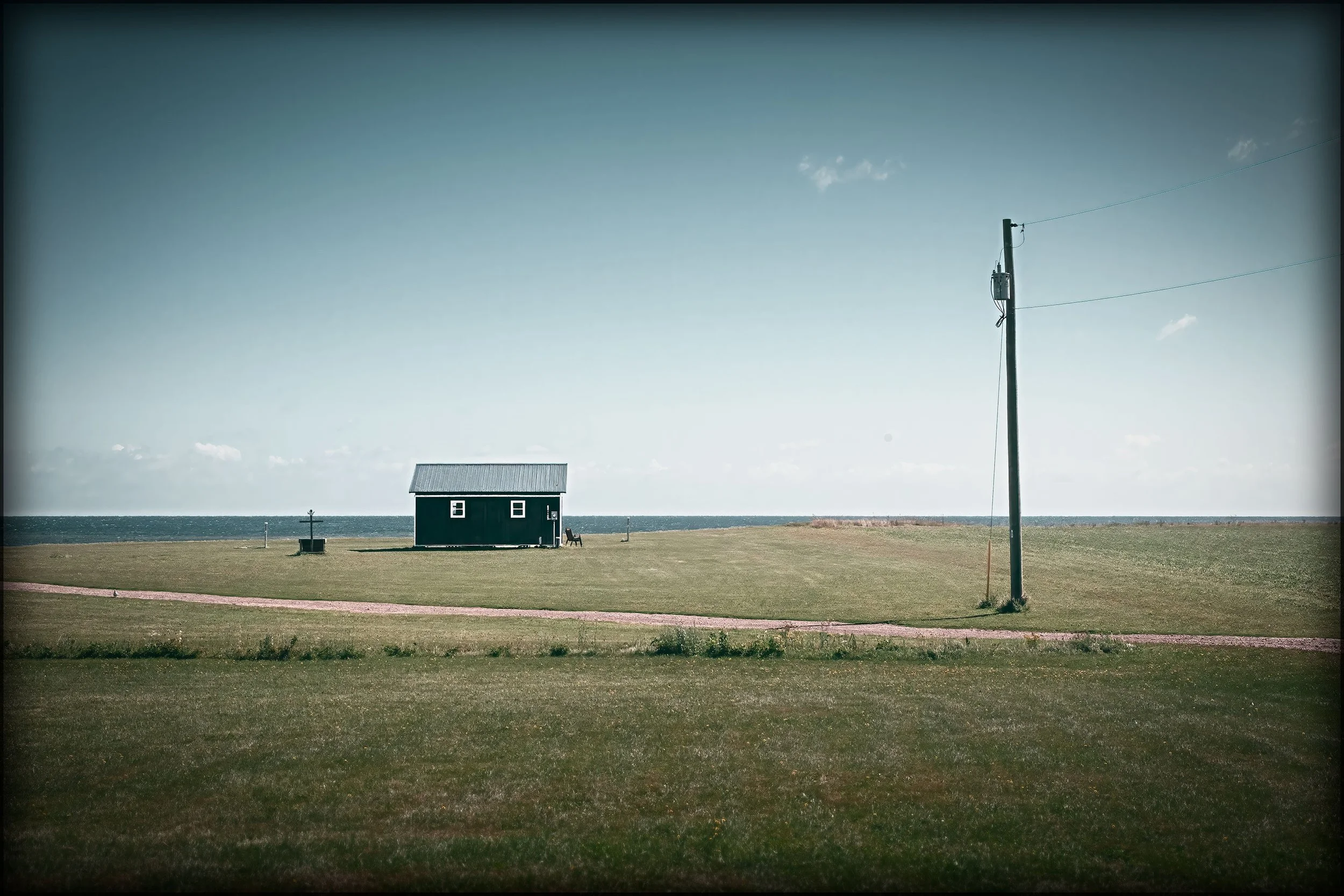 A small dark-colored house with a metal roof is located on a grassy area near the ocean. A utility pole with wires is on the right, and a bench is near the house. The sky is mostly clear with a few clouds.