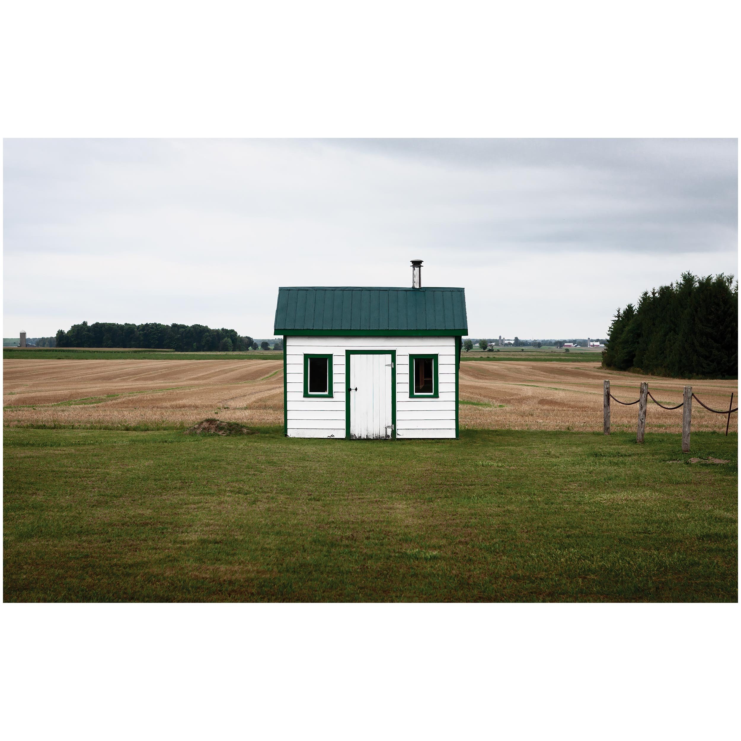 Small white house with green trim and a green roof, sitting in a rural landscape with open fields and trees under a cloudy sky.