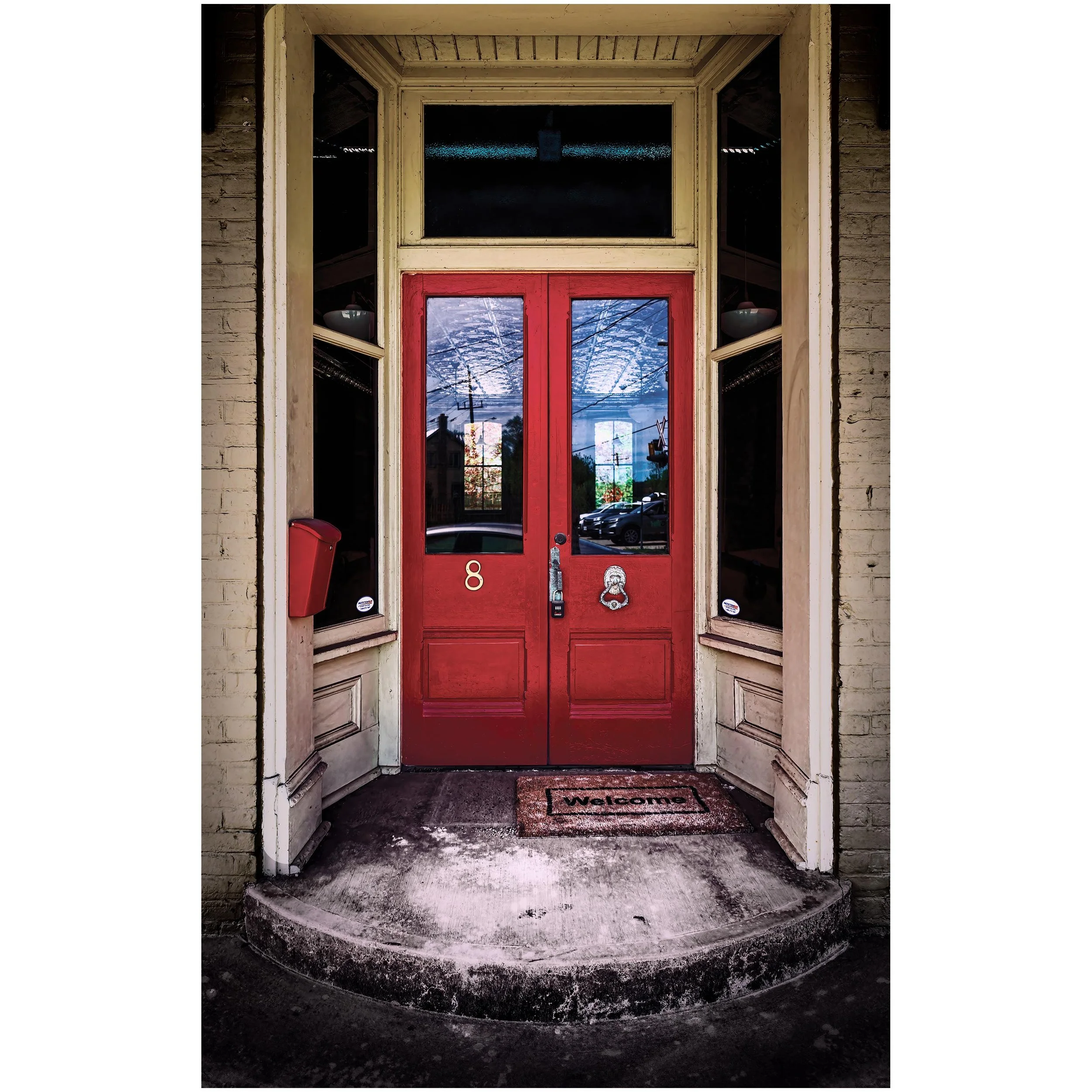 Front entrance of a house with a red door, two glass panels, a BT doorbell, and a barrel-shaped door knocker, with a welcome mat on the concrete step.