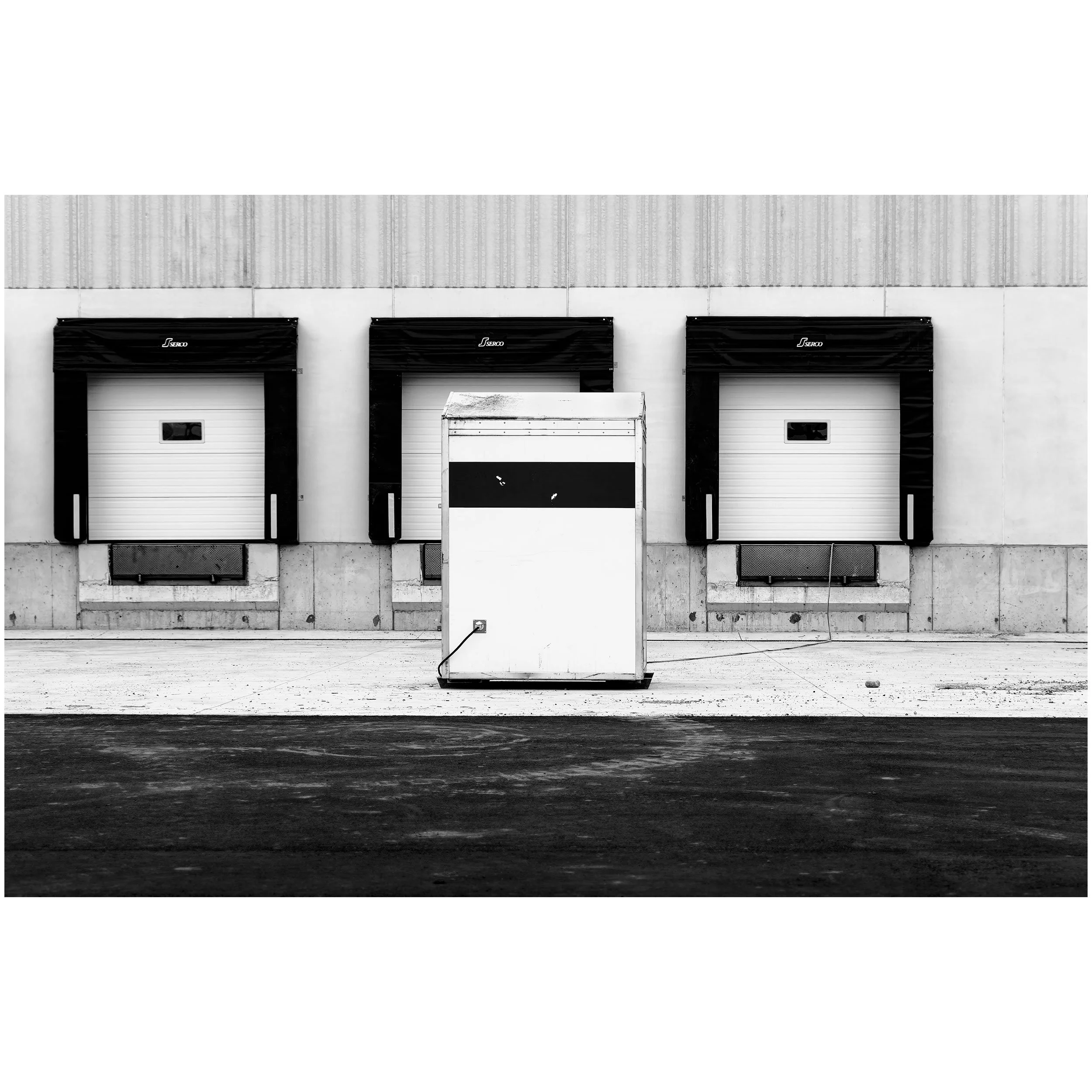 Black and white photo of three large industrial garage doors with black awnings labeled 'Serco' and a white cart in front of the middle door, set against a concrete wall.