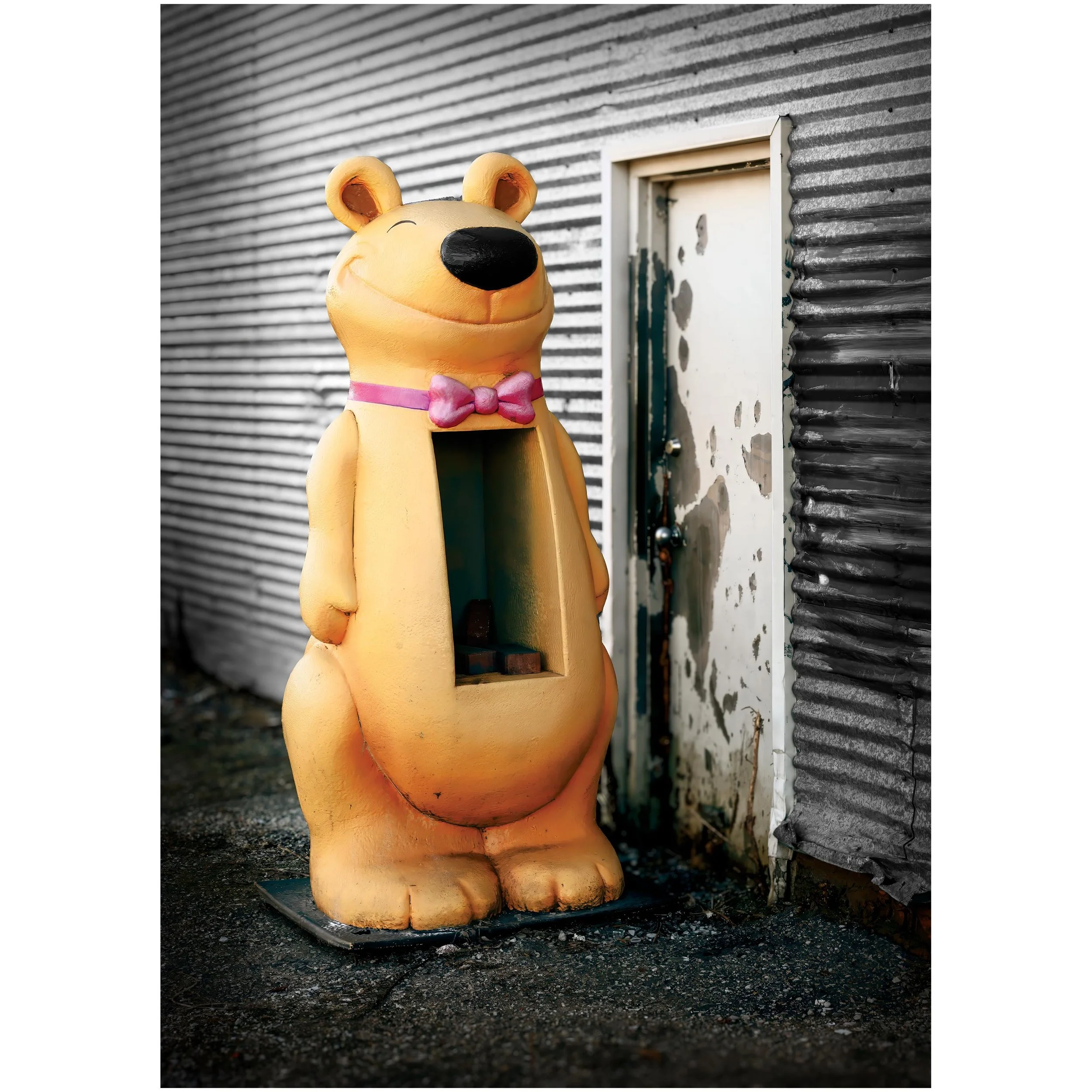 A life sized yellow bear-shaped display with a pink bow sitting on the ground next to a corrugated metal wall and a utility box.