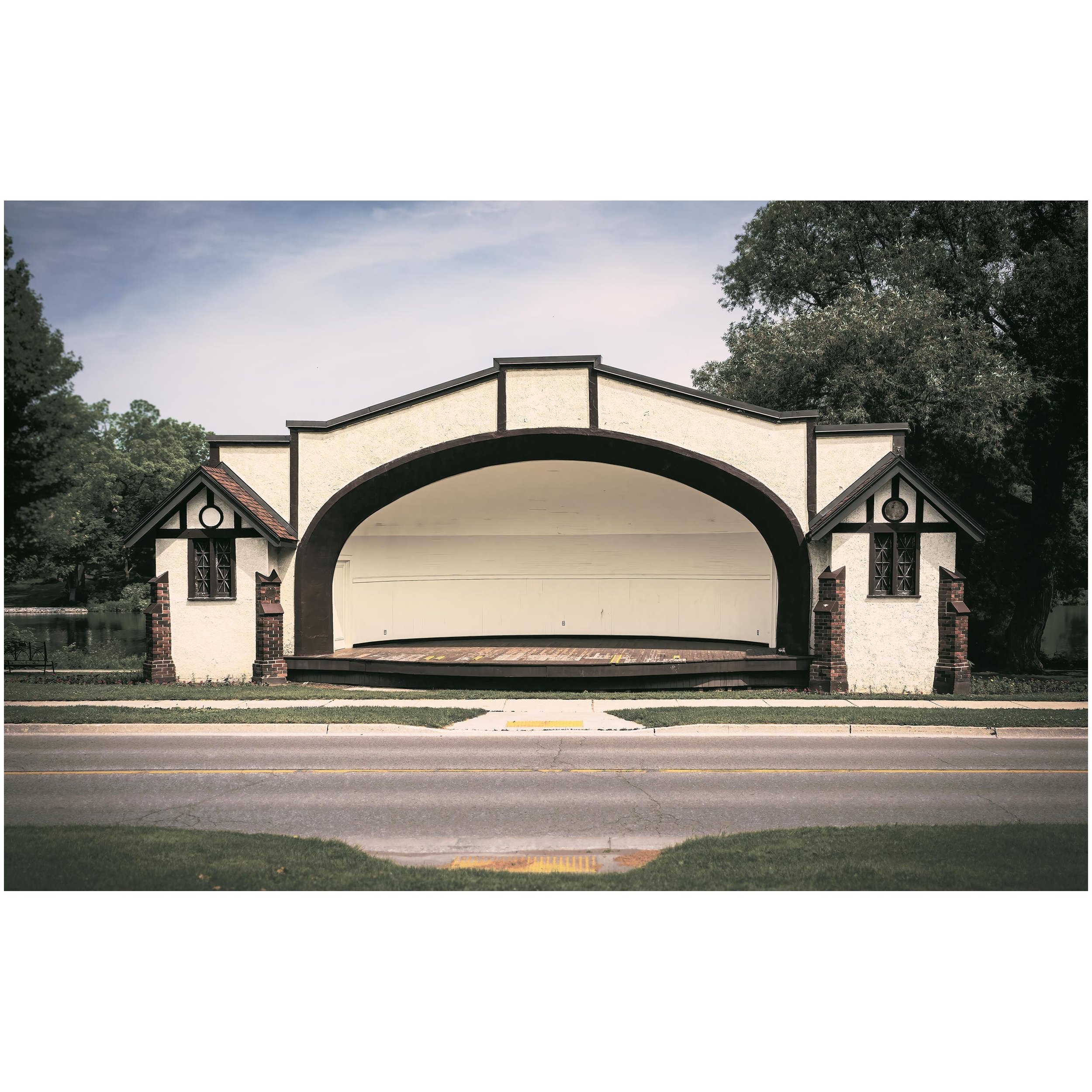 A large shakespearian stage with a large arched opening on a street, surrounded by green trees, under a partly cloudy sky.