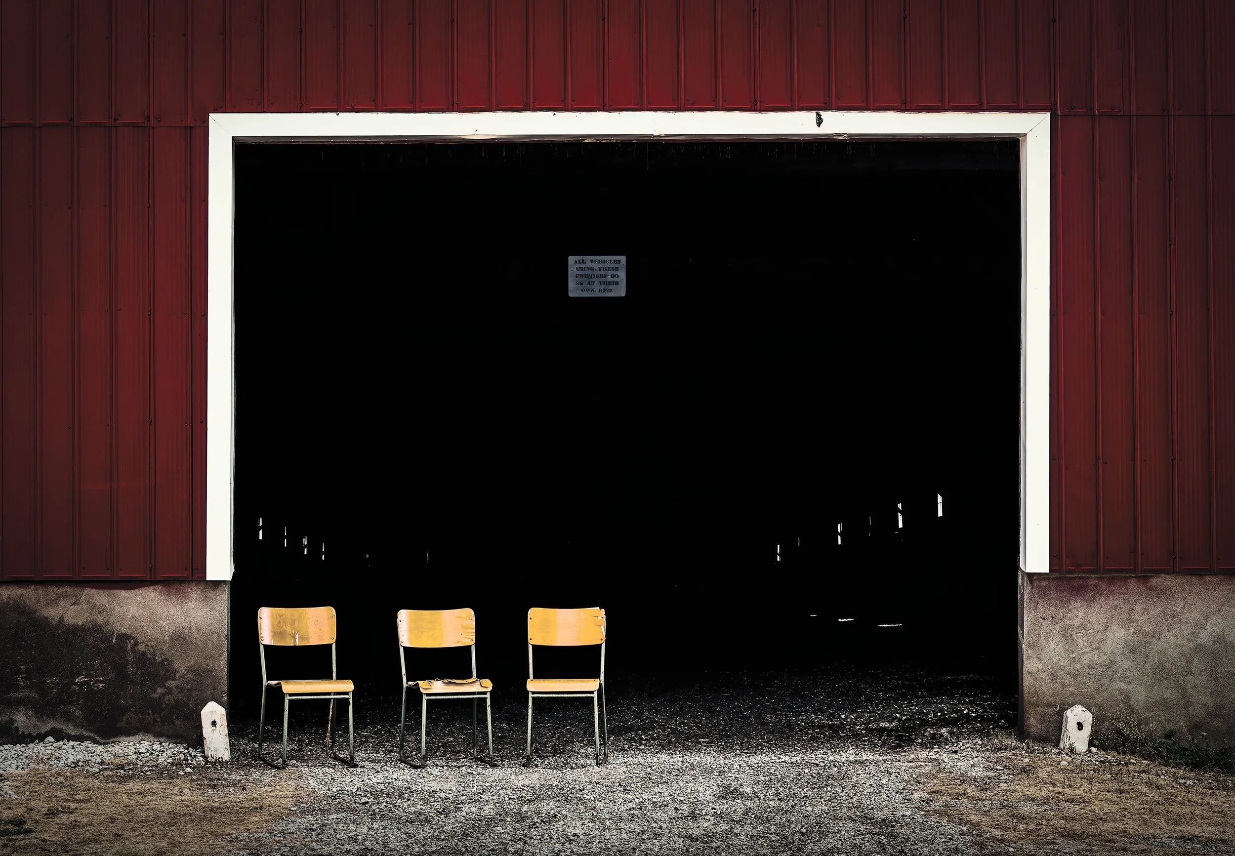 Three yellow chairs with wooden seats and backs are placed outside in front of a dark, open structure with a red exterior wall and a white frame. There is a small sign on the dark structure that reads 'All vehicles using these premises do so at their own risk.'