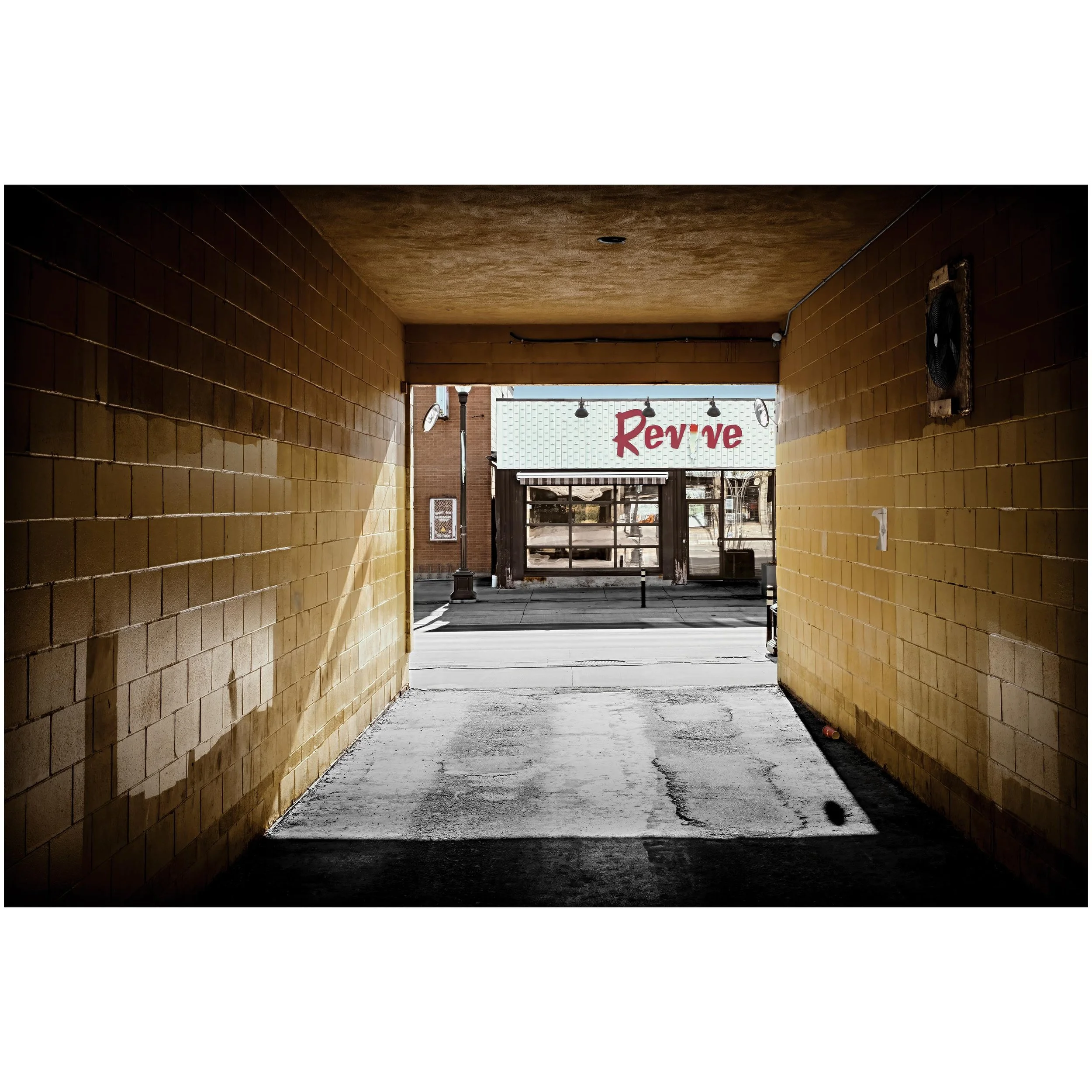 View through a tunnel with brick walls and a yellow ceiling, leading to an open street with a sign that says 'Revive' and street lamps in the background.