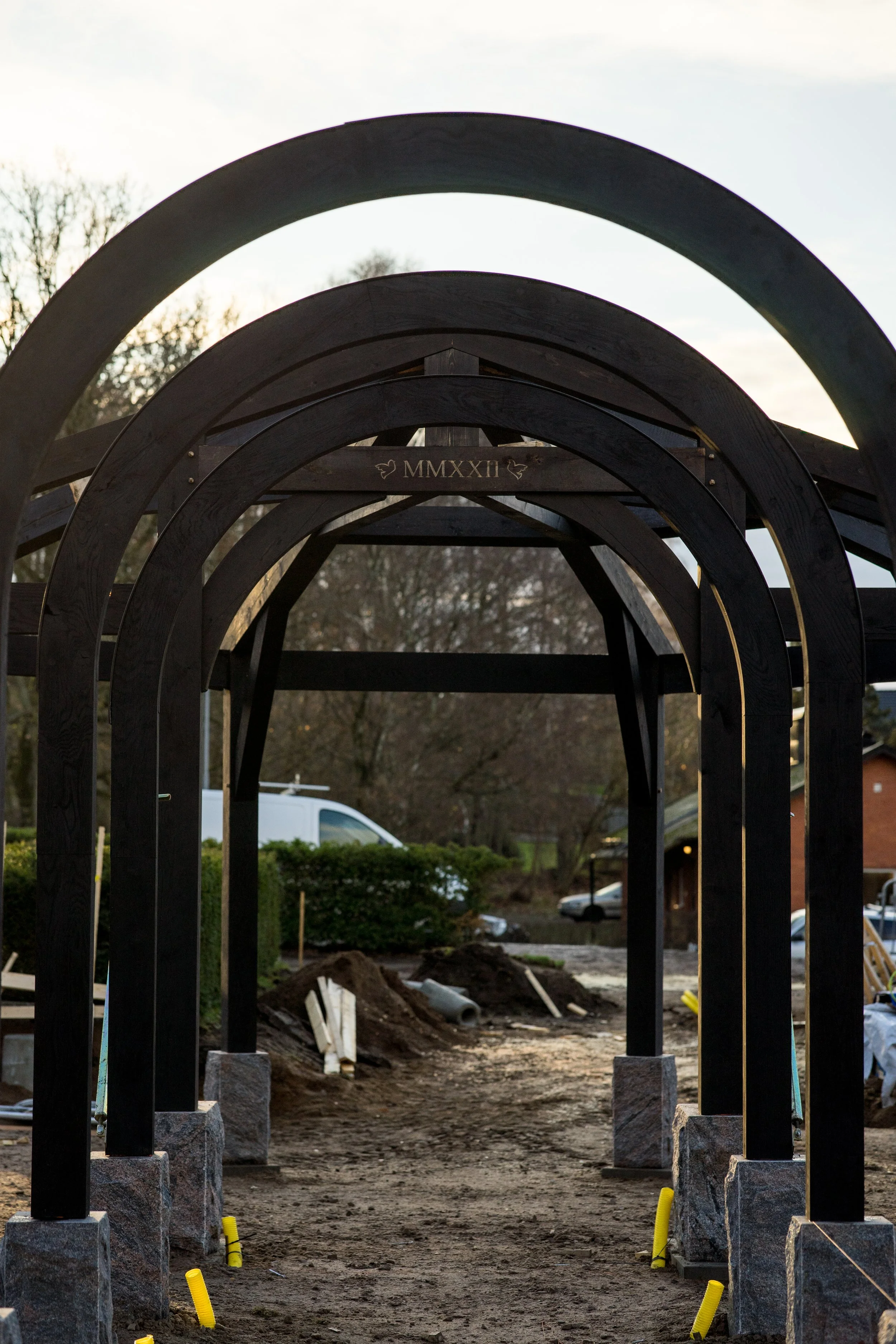 Construction site with a series of dark wooden arches forming a pathway, with trees and vehicles in the background, and the Roman numerals MMXXII engraved on the top arch.