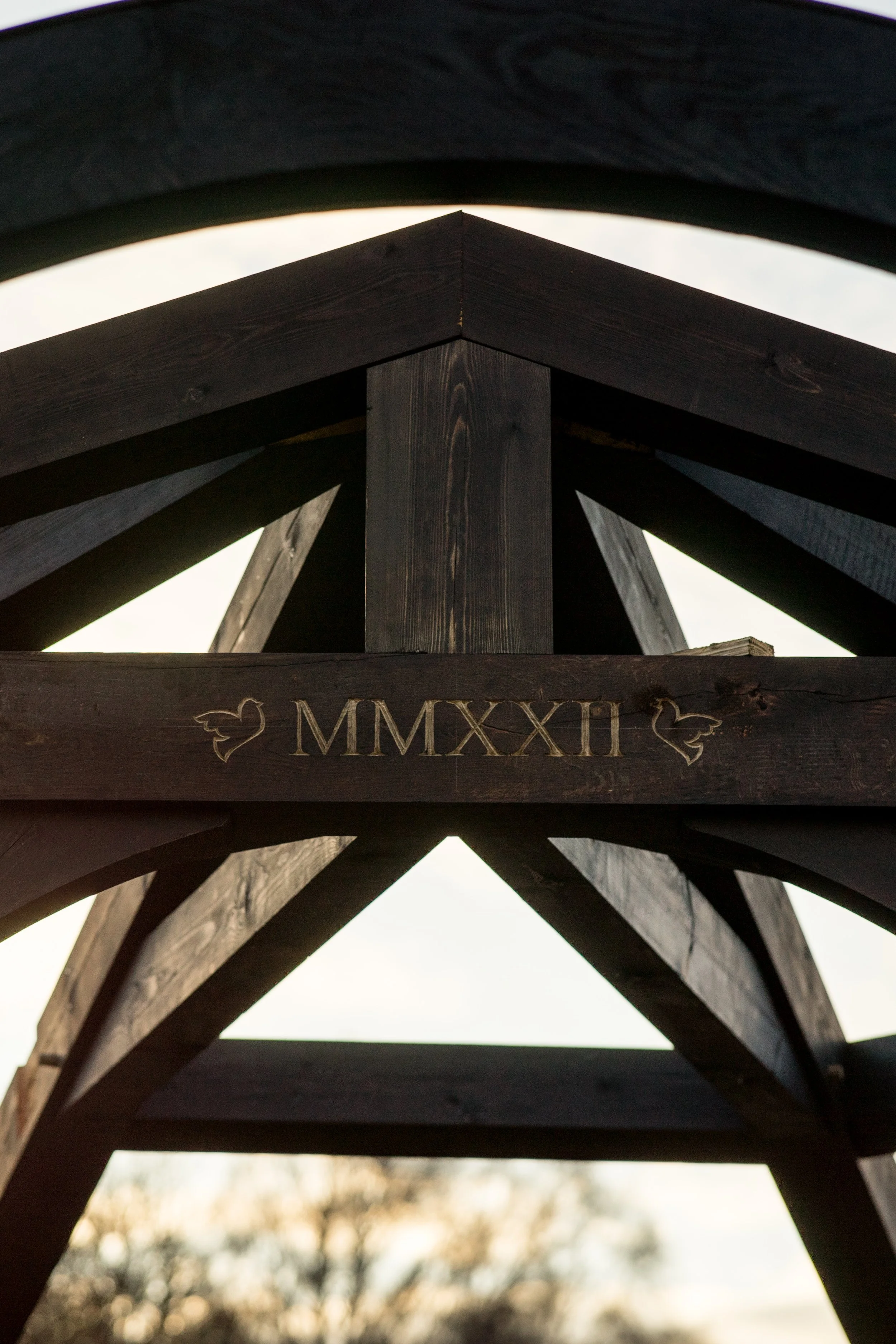 Looking up at a wooden structure with engraved Roman numerals 'MMXXII' and decorative birds, with a cloudy sky and trees in the background.
