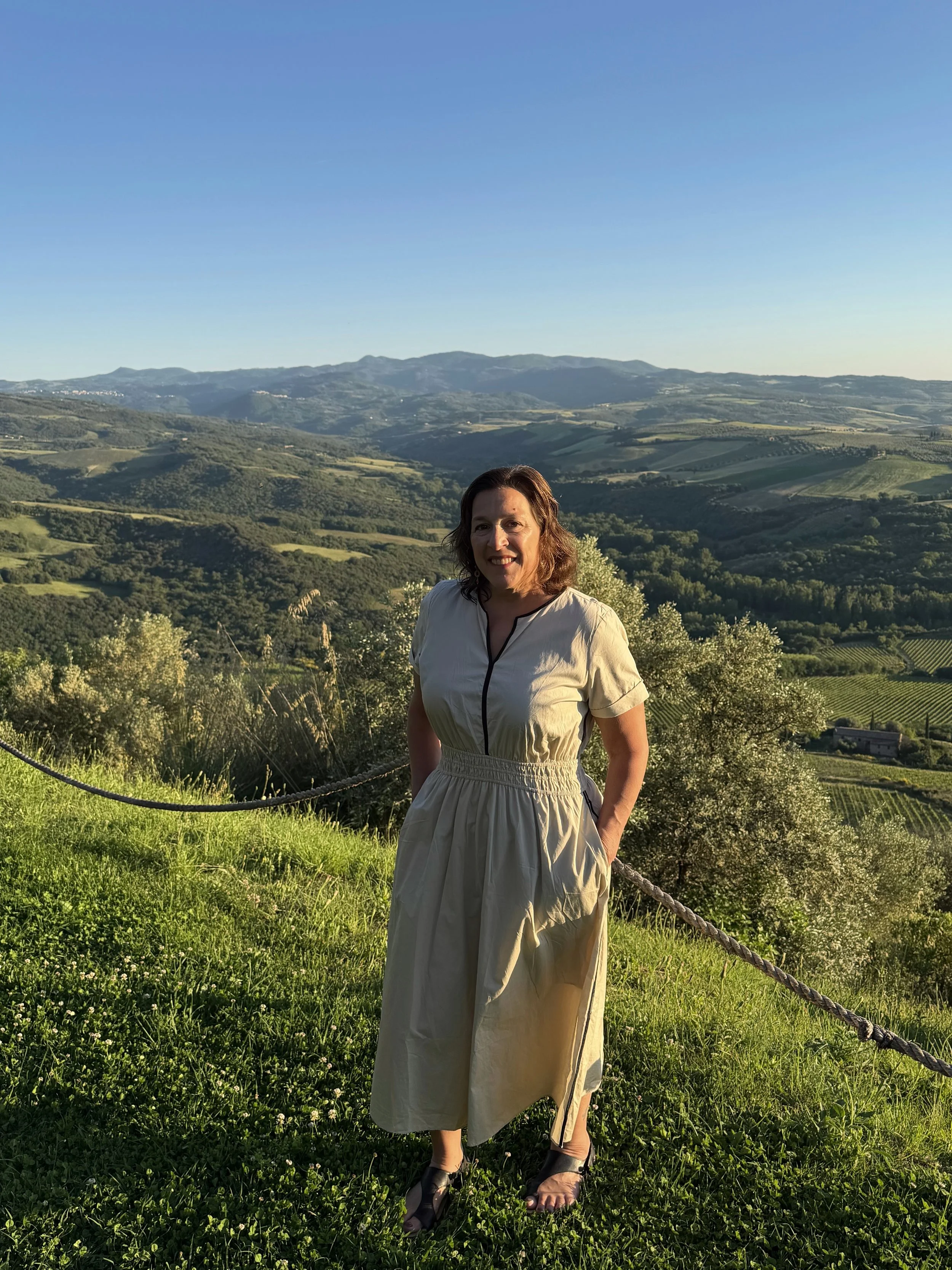 Woman standing outdoors on green grass with a scenic view of rolling hills and mountains in the background during sunset.