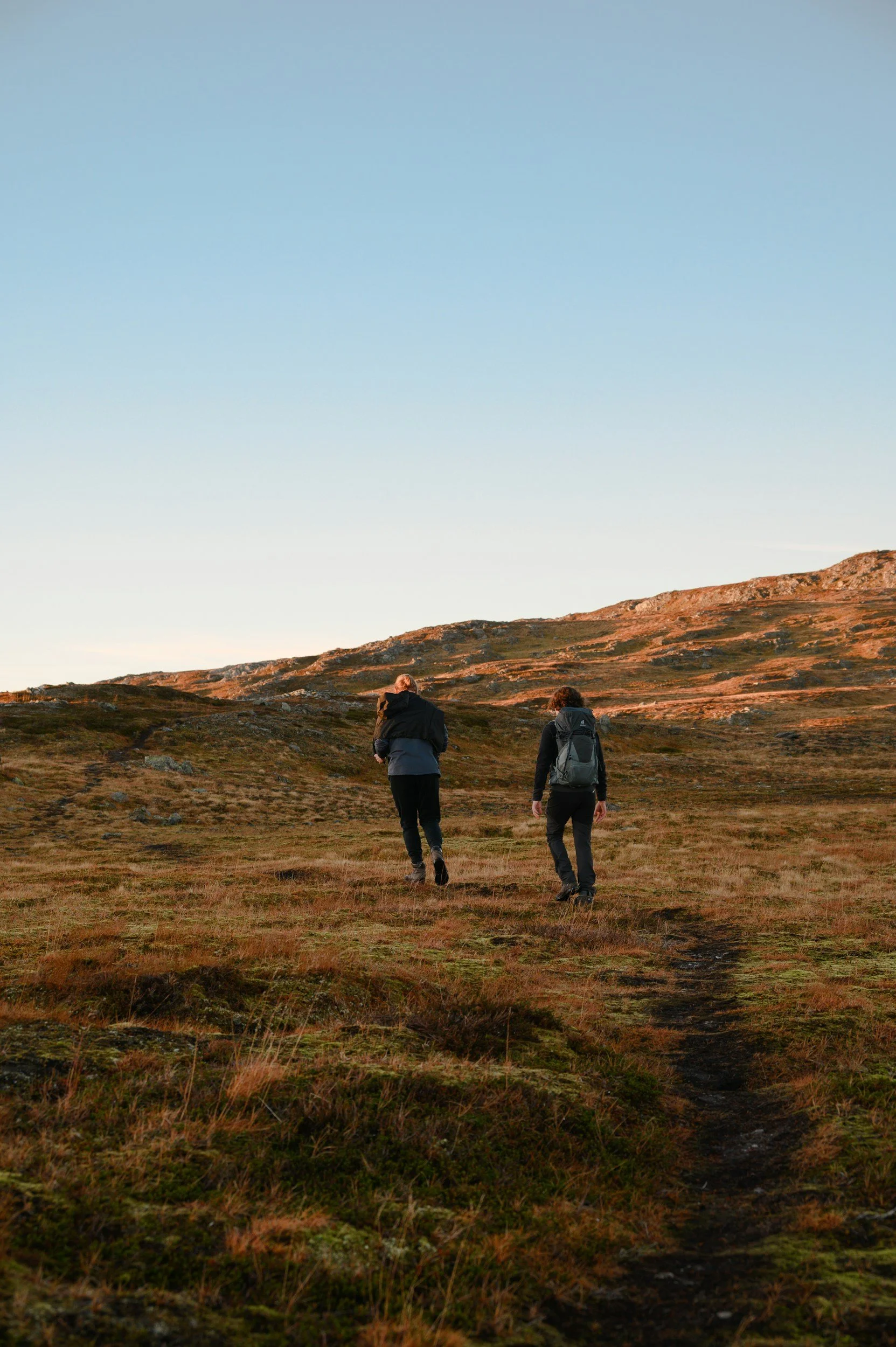 Two hikers walking on a trail through a grassy landscape with hills under a clear sky.