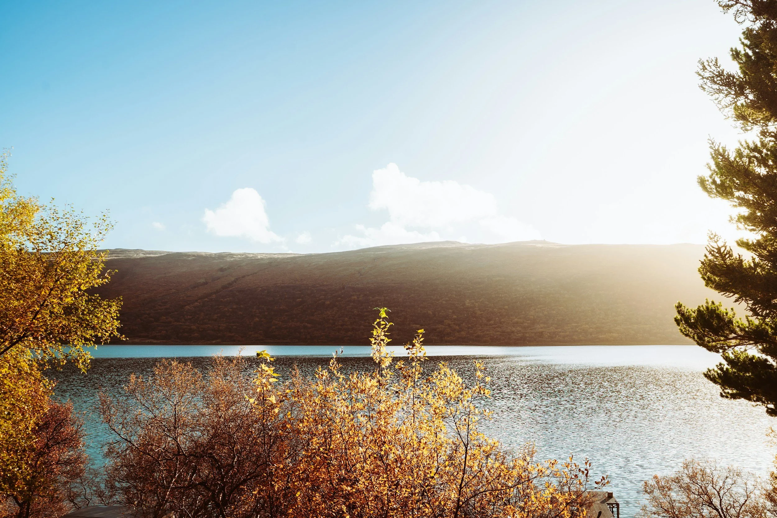 A scenic view of a lake with calm water surrounded by autumn foliage, a hillside in the background, and trees on the right and bottom edges of the image, bathed in warm sunlight.