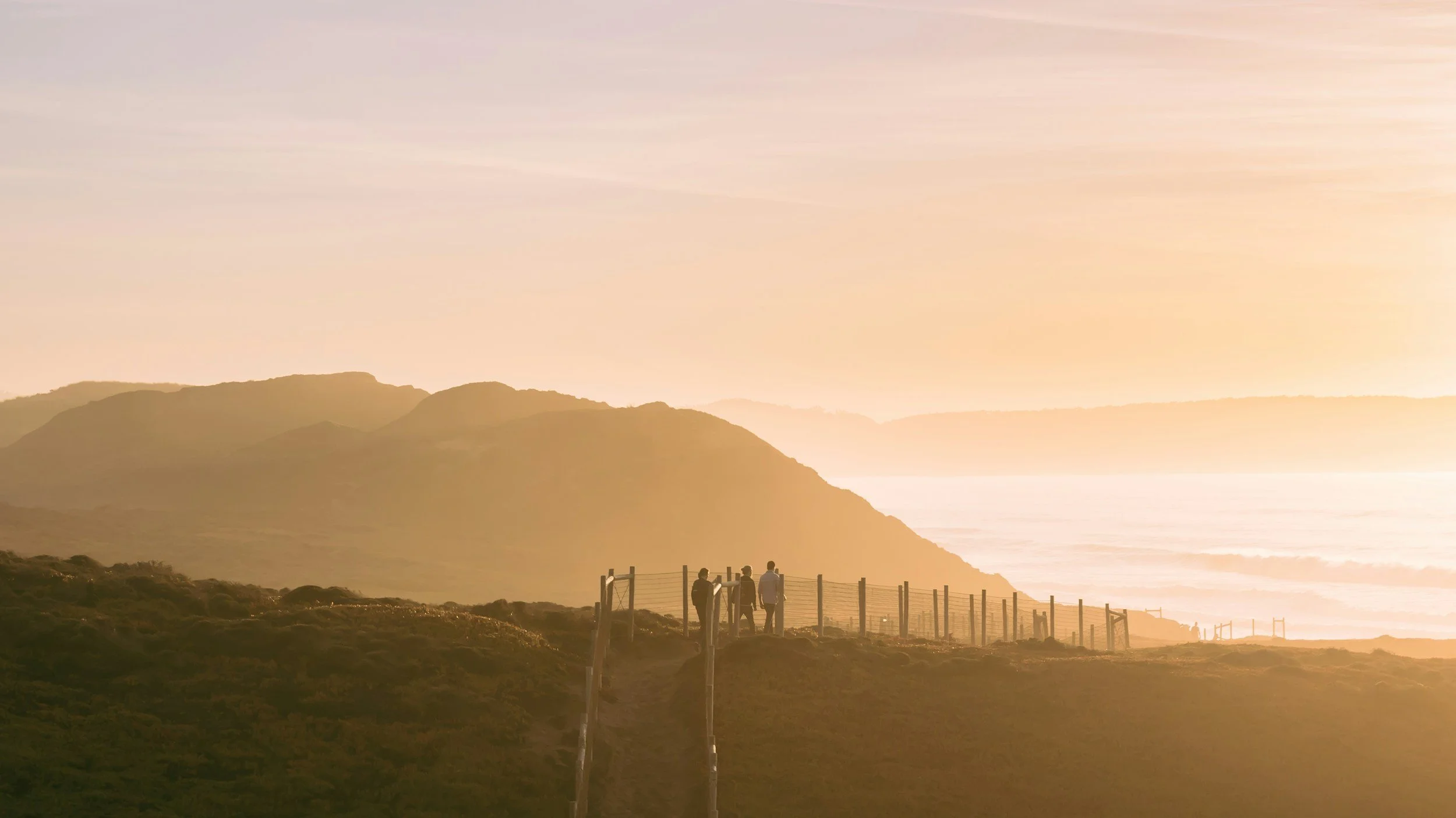 Four people standing near a fence on a hilltop overlooking a coastal landscape at sunset, with mountains and the ocean in the background.