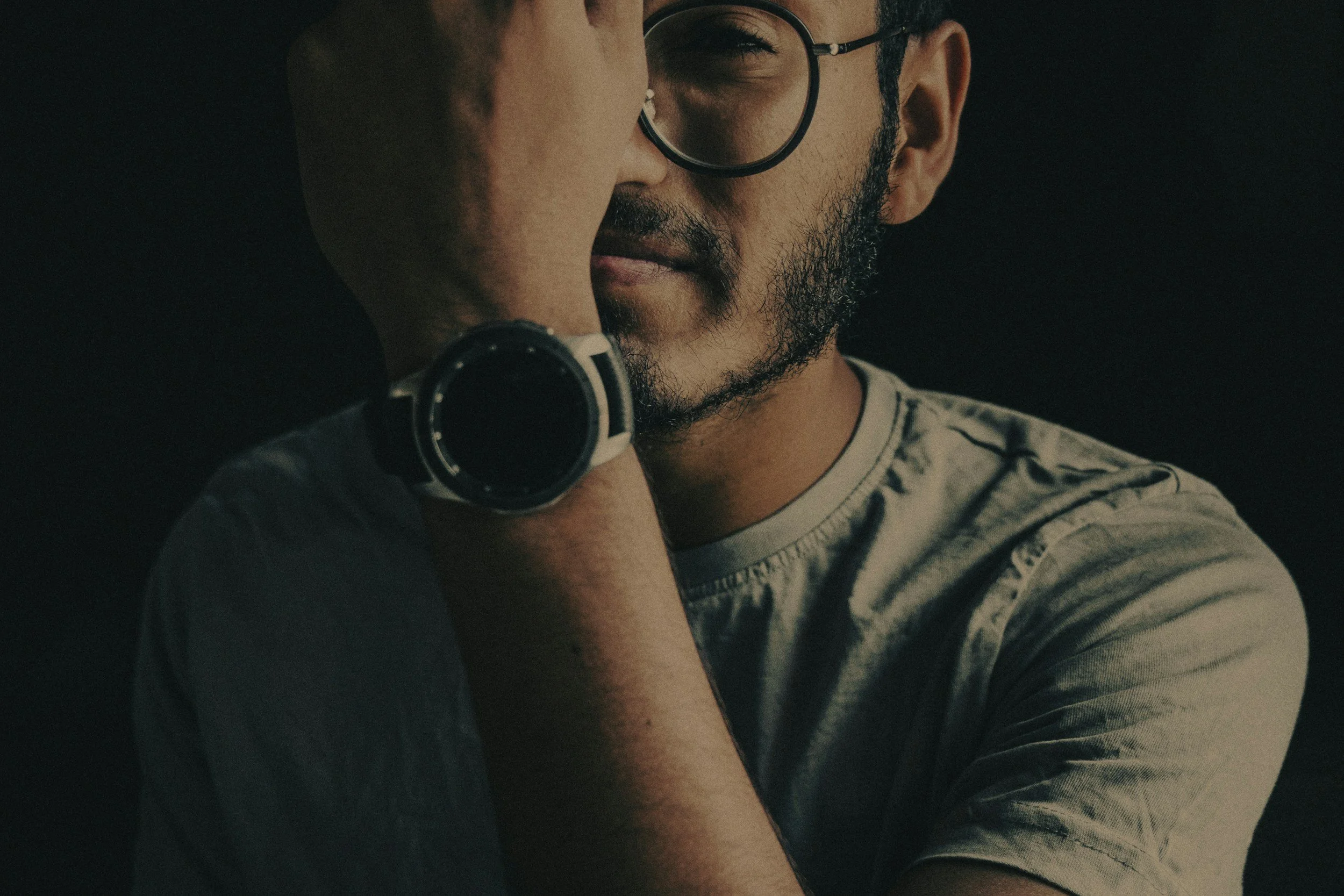 A close-up of a man with glasses and a beard, whispering into a woman’s ear, who is partially visible.