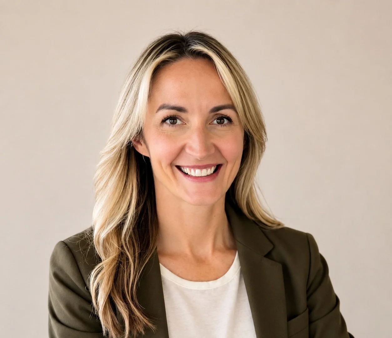 Headshot of a smiling woman with blonde hair, wearing a dark blazer and white top, standing against a plain background.