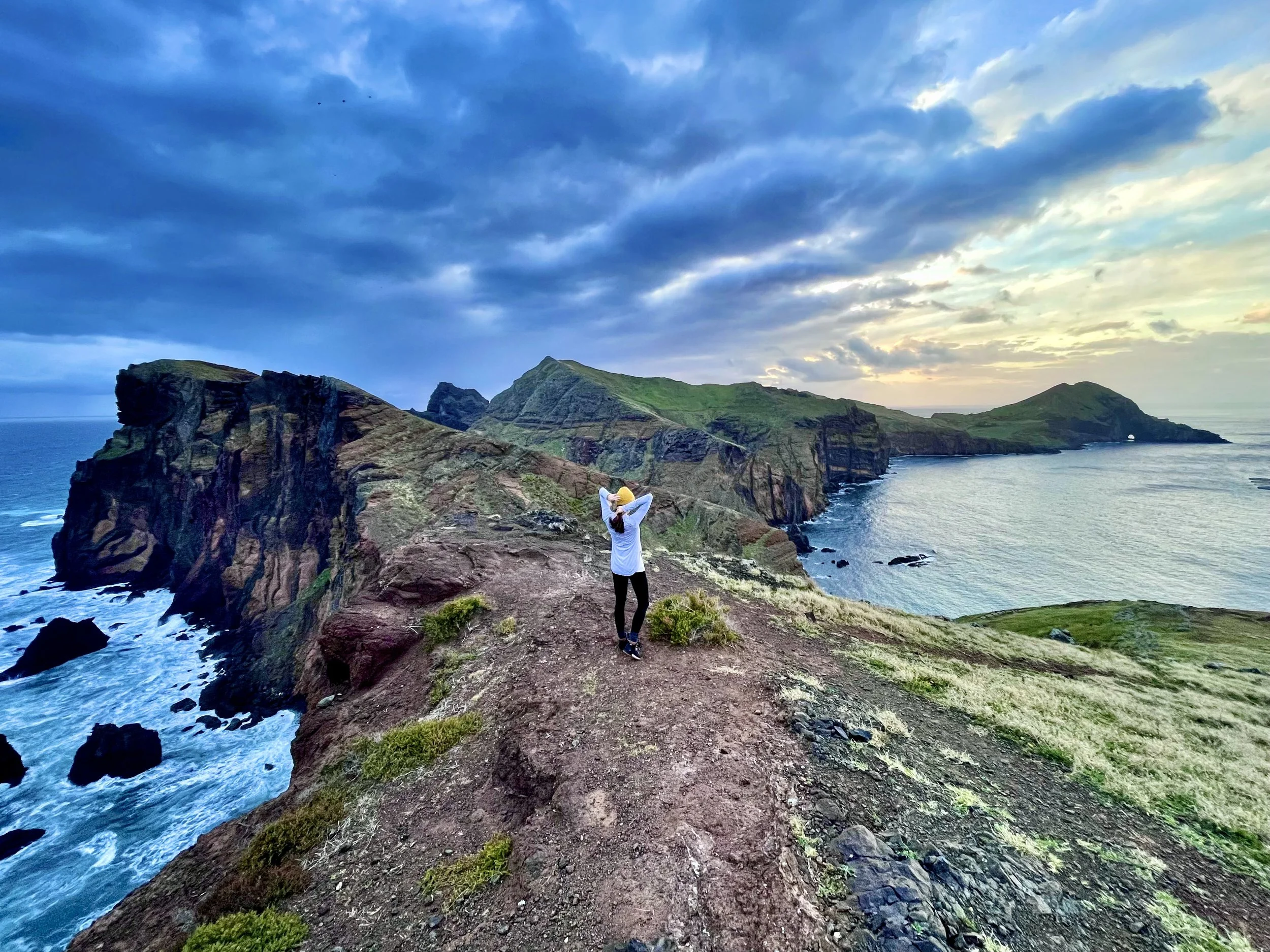 Person standing on a cliff overlooking a rugged coastline with cliffs, ocean waves, and a partly cloudy sky at sunset or sunrise.