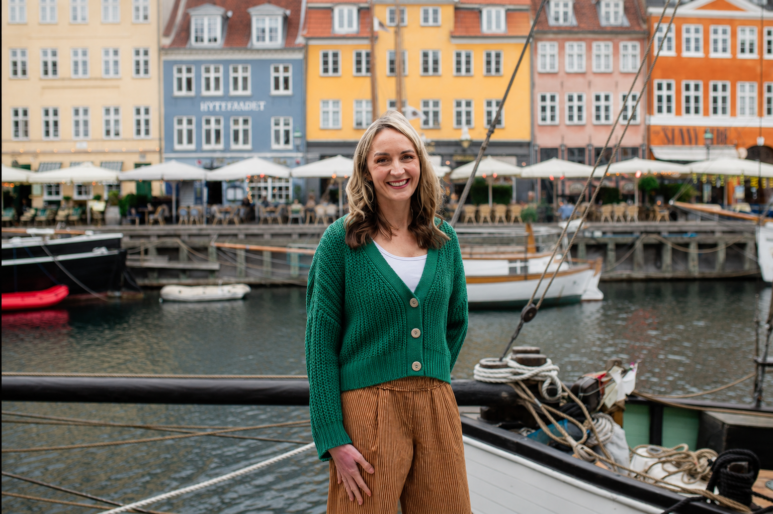 A woman with shoulder-length wavy brown hair smiling and wearing a green cardigan and brown pants, standing near a harbor with boats and colorful buildings in the background.