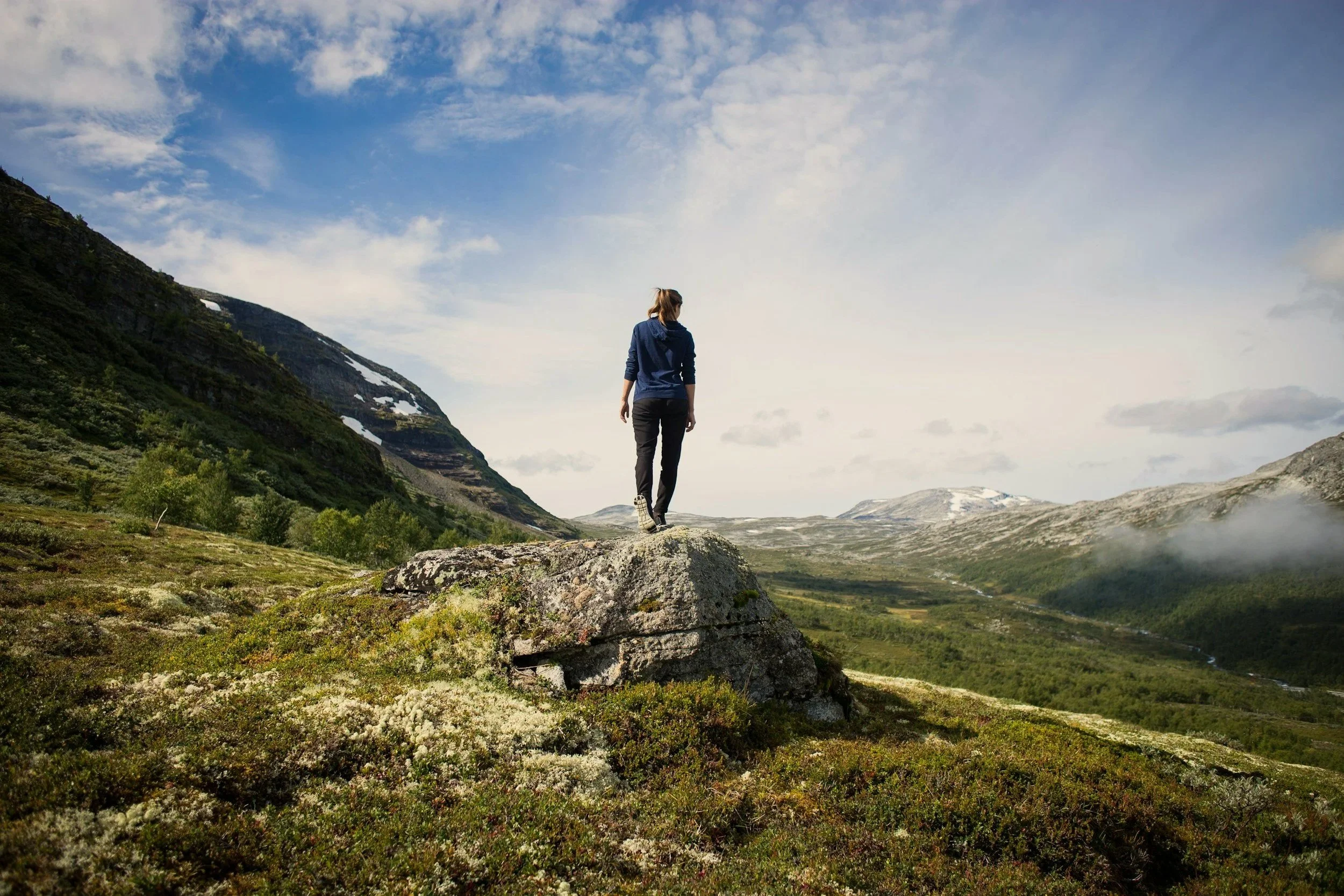 A person standing on a large rock in a lush valley with mountains in the background and a partly cloudy sky.