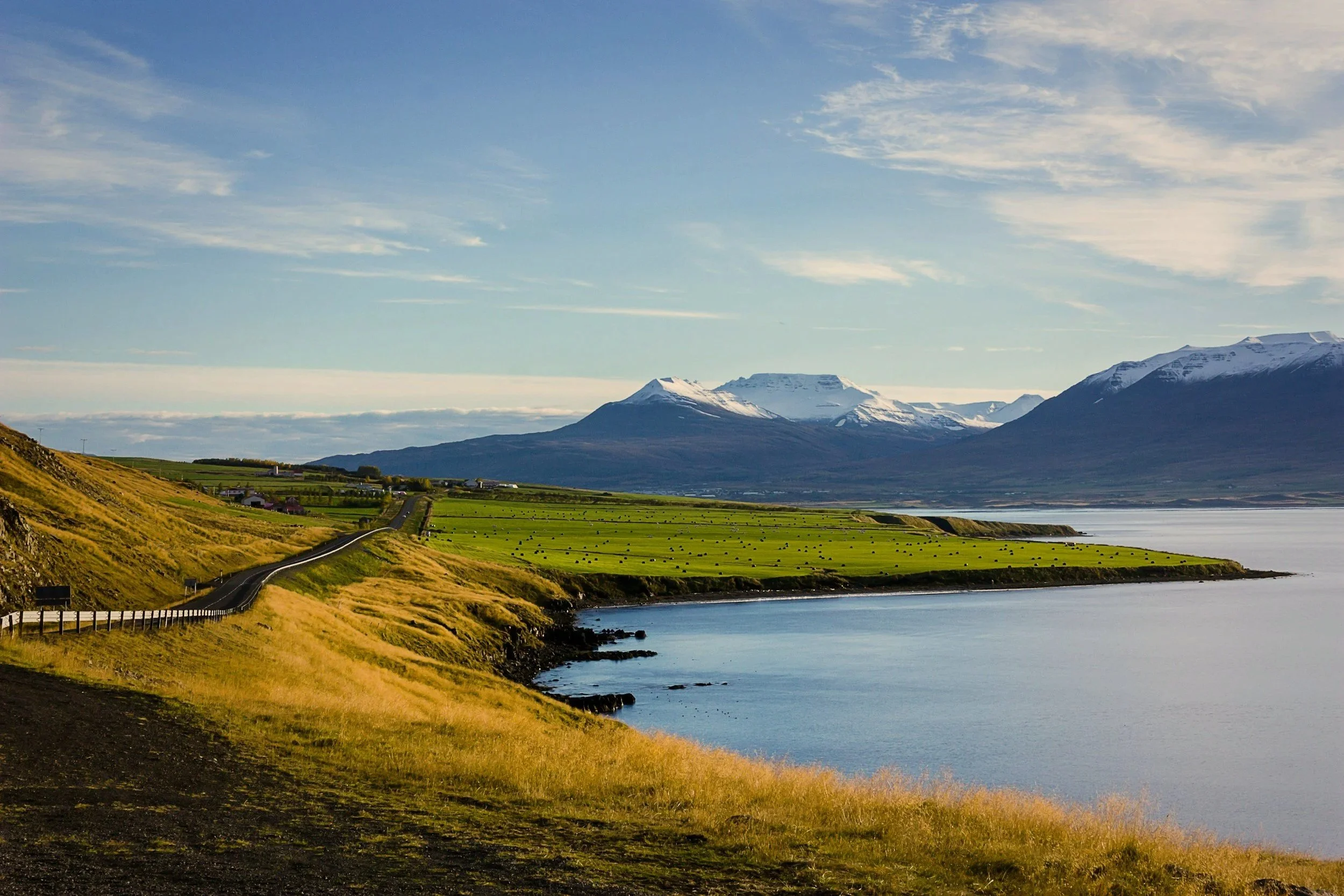 Scenic view of a calm body of water with rolling green fields and a winding road along the shores, mountains in the distance with snow-capped peaks, and a clear blue sky with some clouds.