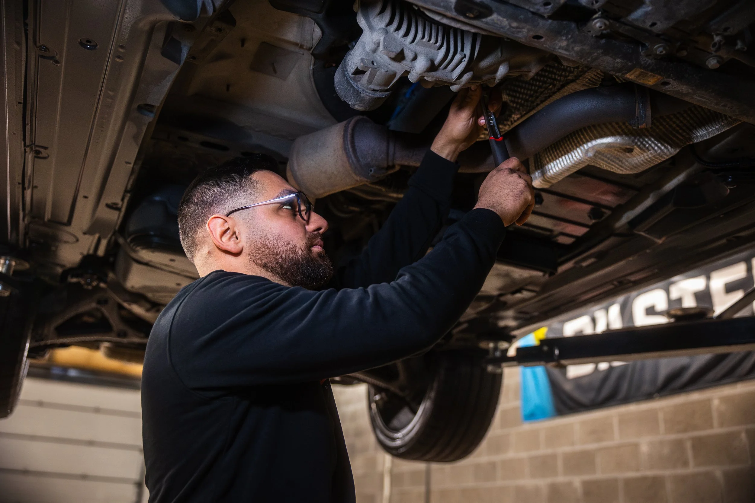 Un homme en train de réparer une voiture dans un atelier à Verviers, utilisant un outil pour travailler sous le véhicule.