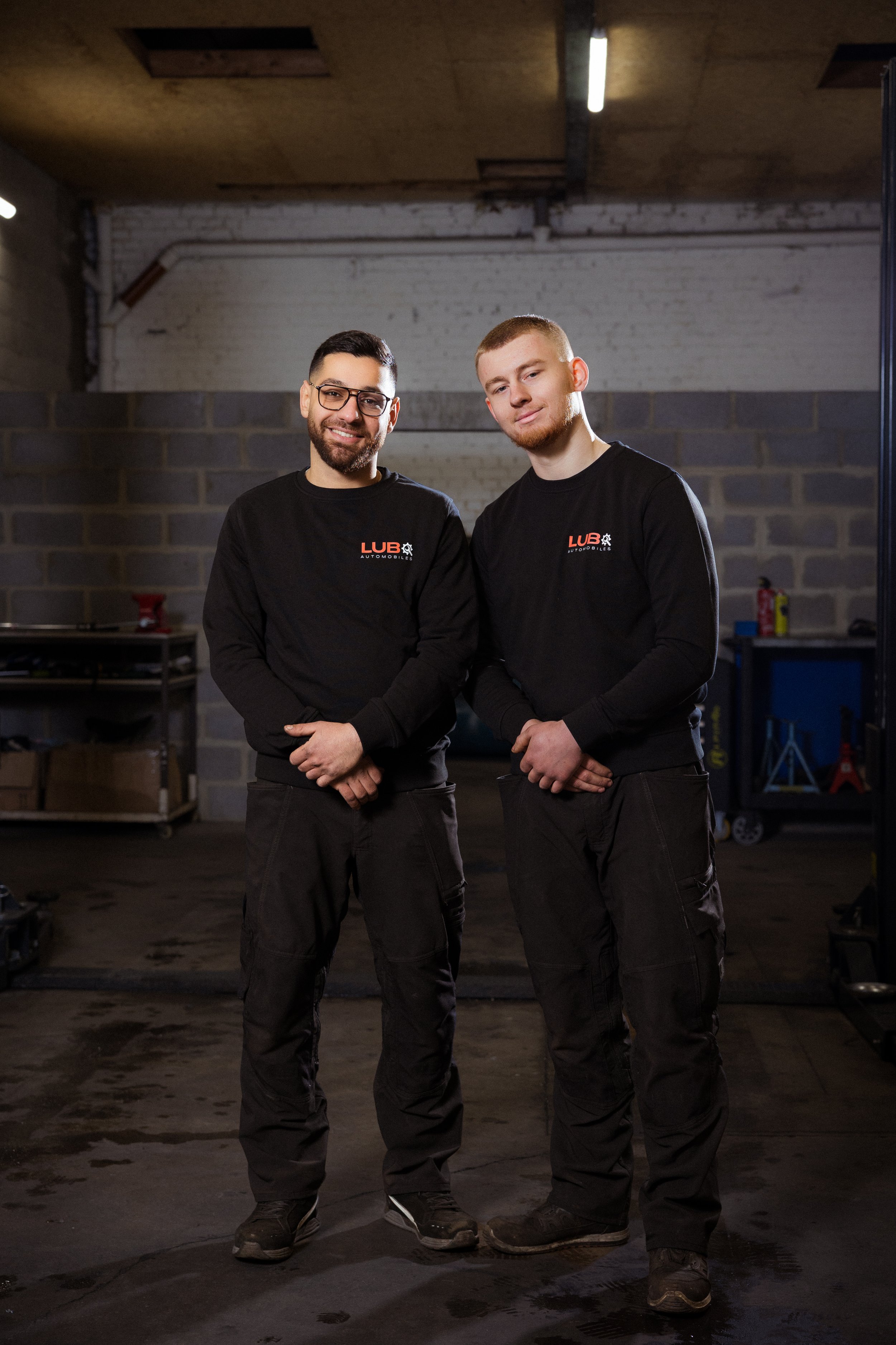 Deux hommes en vêtements de travail noir avec le logo LUBO Automobiles, debout dans un atelier à Verviers, souriant à la caméra.