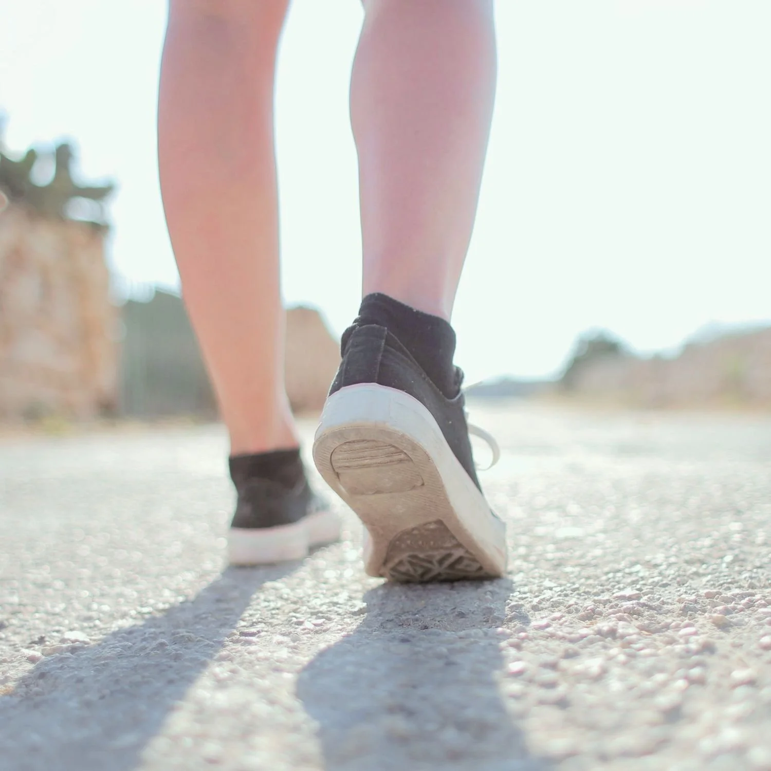 Close-up of a person walking on a gravel path wearing black sneakers and black socks, with the sunlight casting a shadow on the ground.