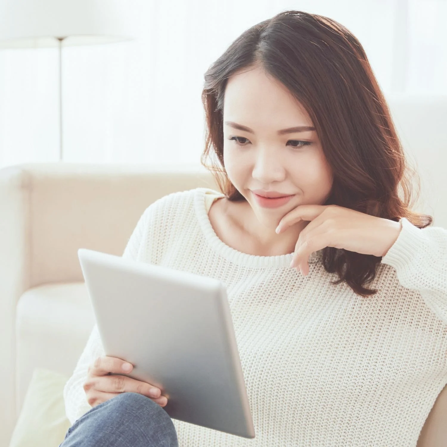 Young woman sitting on a beige couch using a tablet in bright living room.