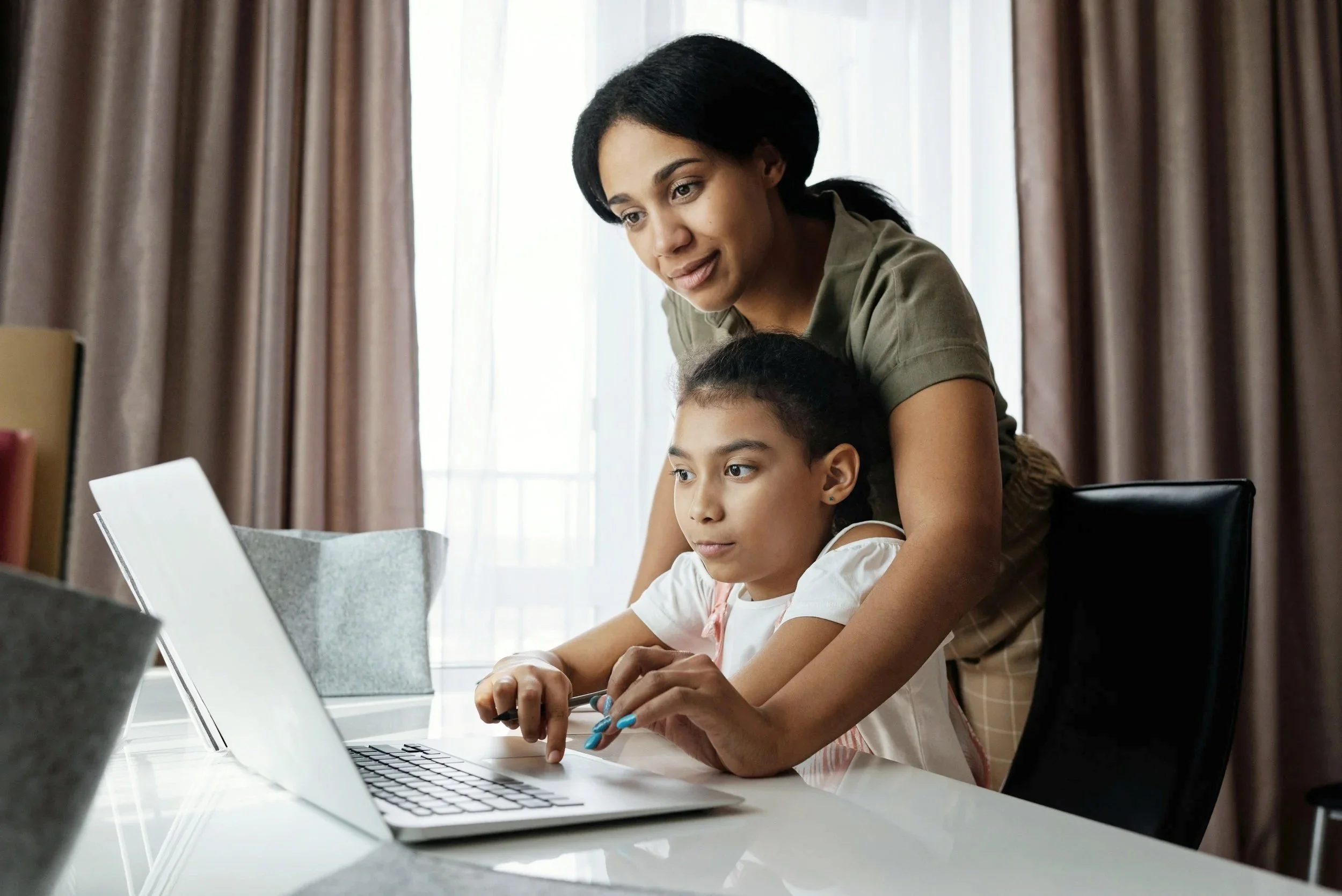 A woman help a young girl using a laptop at a desk in a room with closed curtains.
