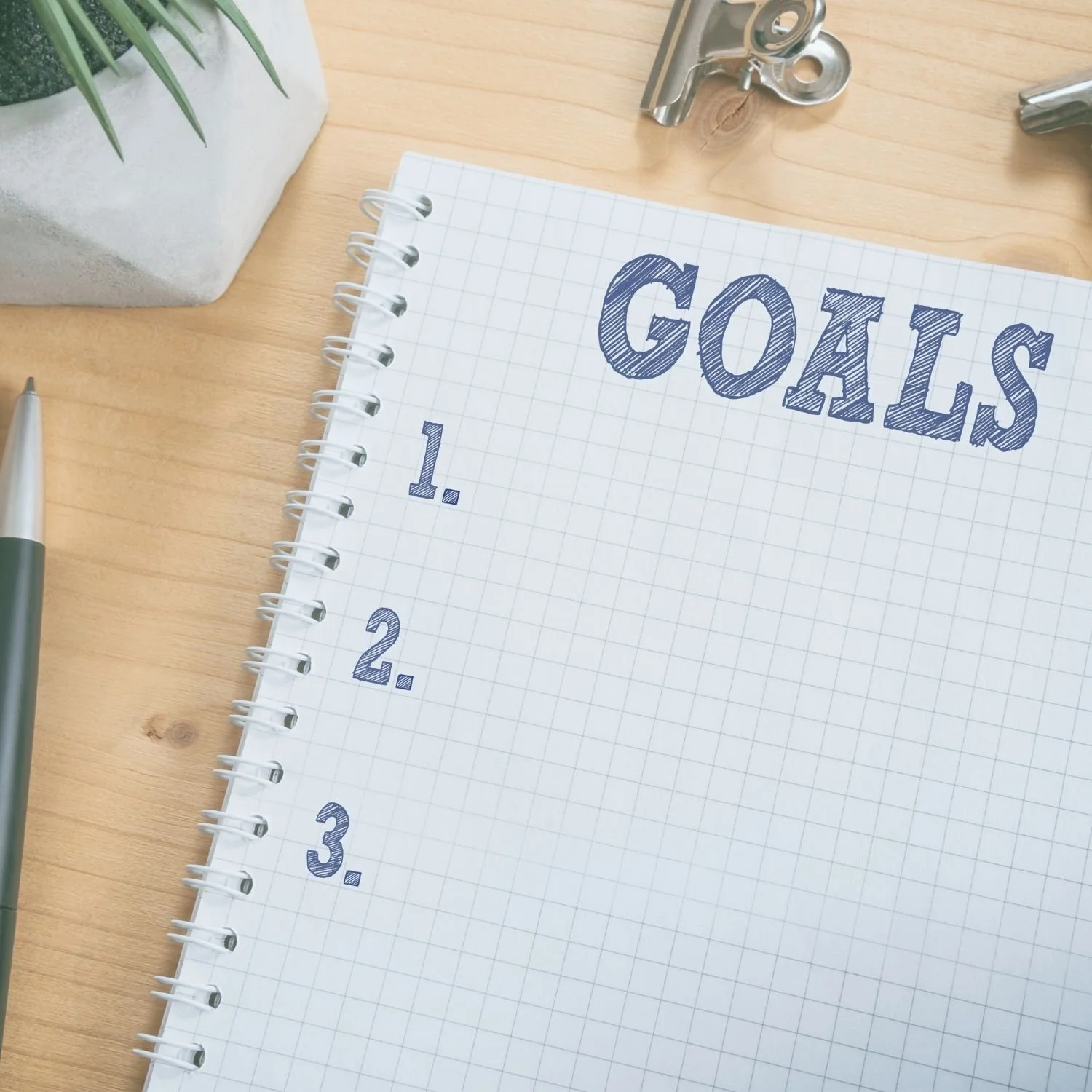 A notebook with 'Goals' written at the top, numbered 1 to 3, on a wooden desk with a pen, a potted plant, and some metal tools nearby.