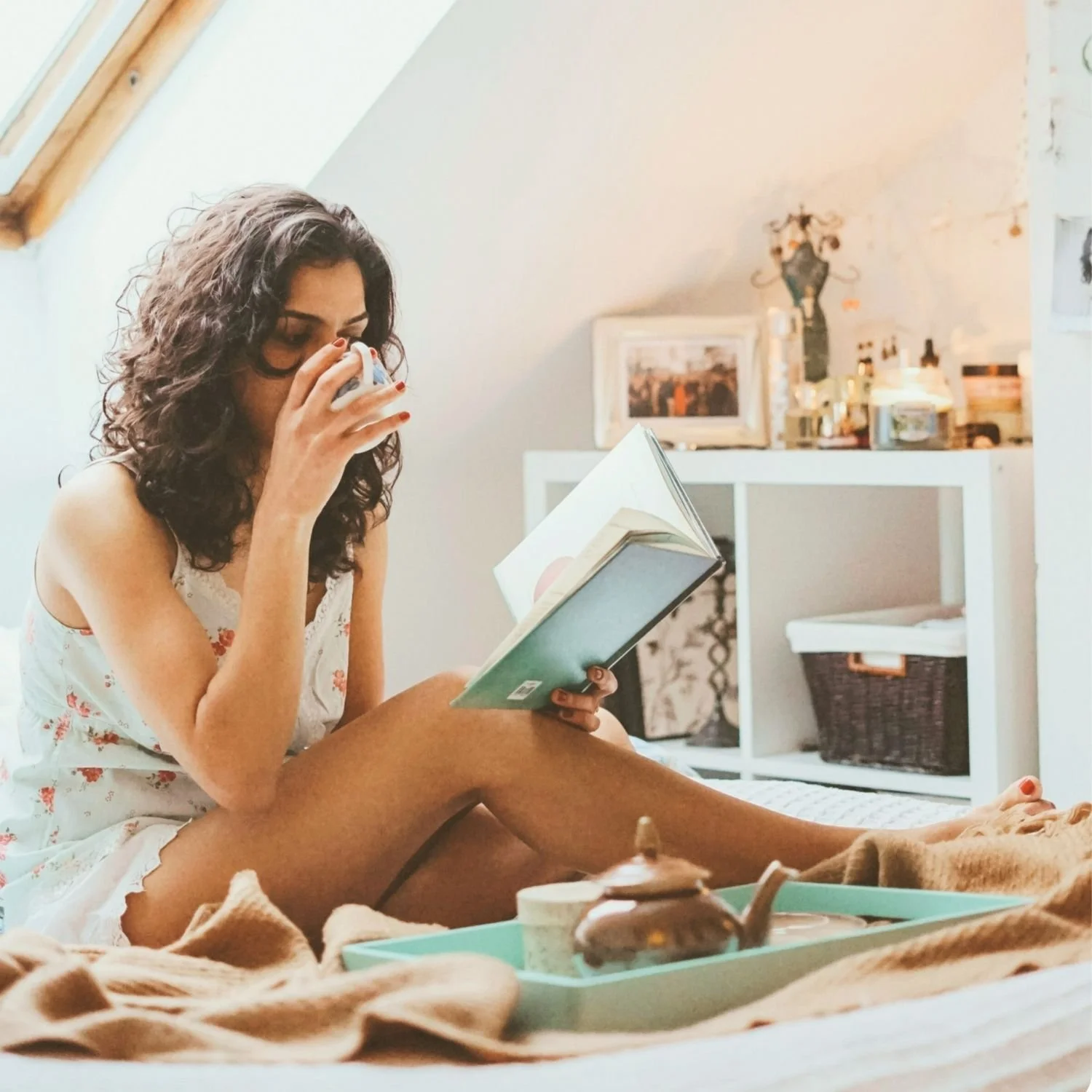 Woman sitting on bed with a book, drinking from a cup, in a cozy bedroom setting with a tray, teapot, and blanket.