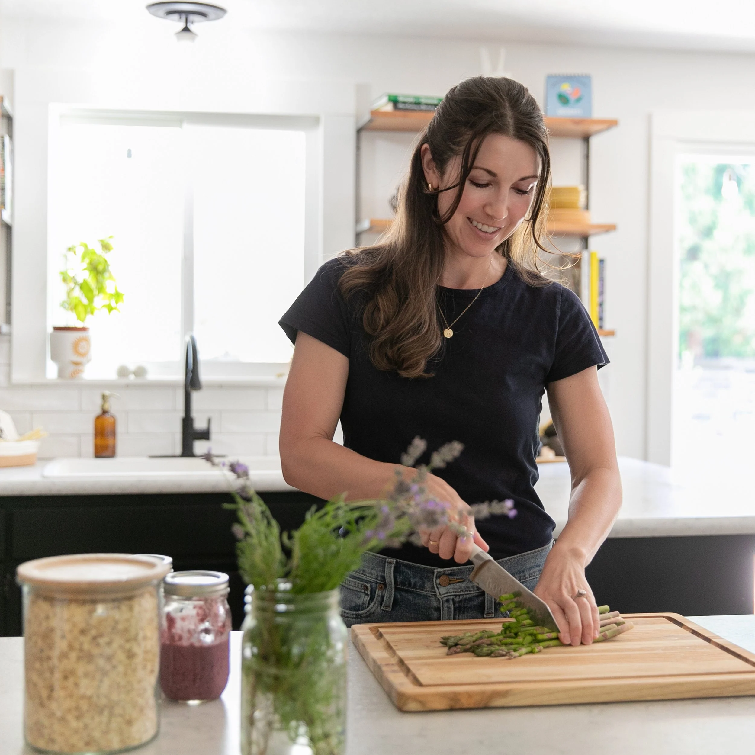 A woman, Ashley Thunell, with long brown hair, wearing a black t-shirt and jeans, is chopping green asparagus on a wooden cutting board in a bright, modern kitchen.