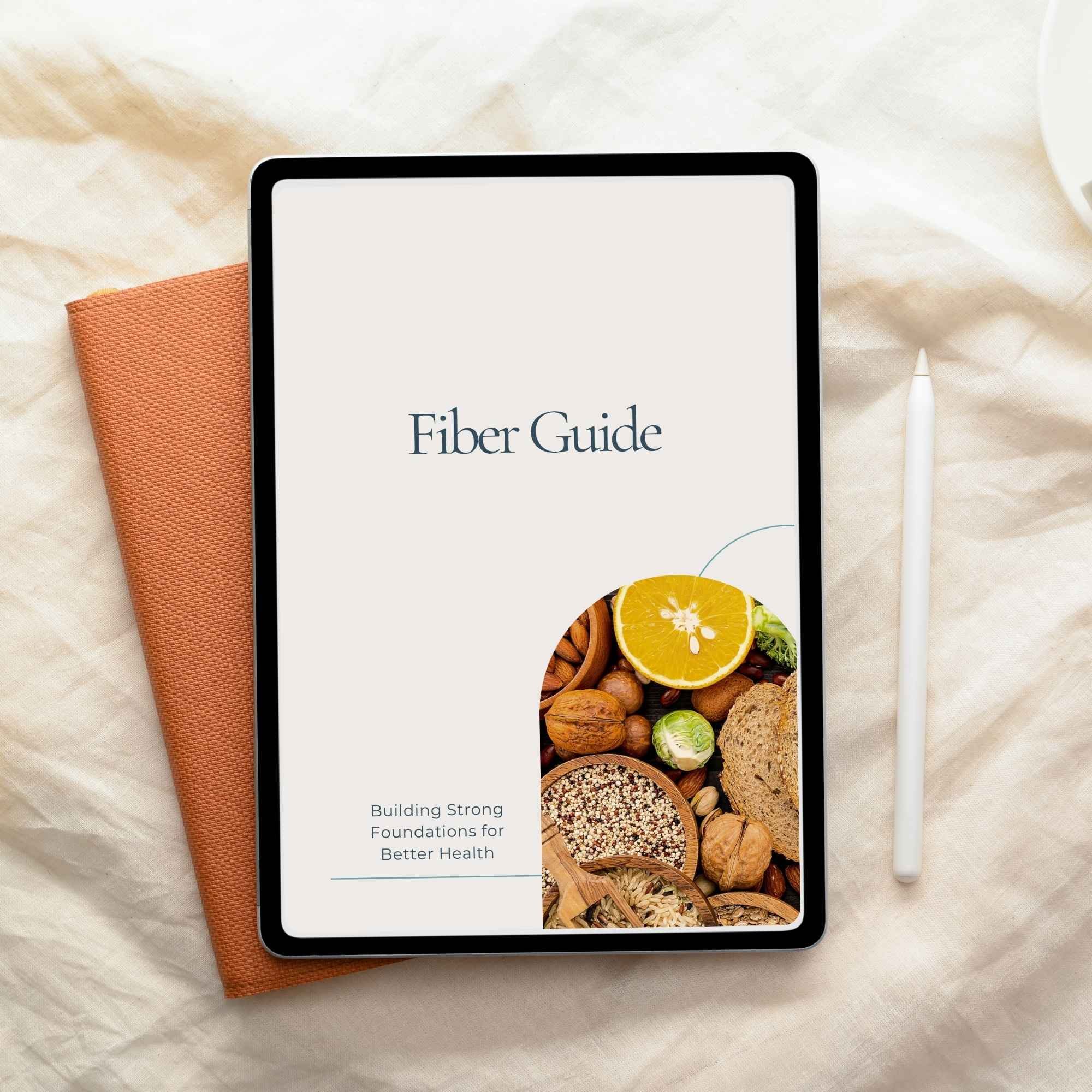 Digital tablet displaying a cover titled "Fiber Guide" with a subtitle "Building Strong Foundations for Better Health" and an image of assorted nuts, seeds, an orange slice, and bread on the screen, placed on a beige fabric surface with a white stylus pen and an orange textured notebook nearby.