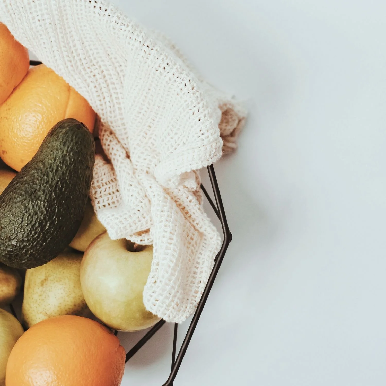 A metal wire basket filled with oranges, pears, an avocado, and a white mesh cloth against a plain light background.