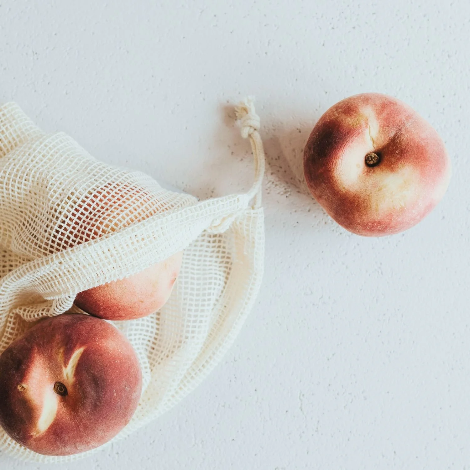 Three peaches and a reusable mesh bag on a white textured surface.
