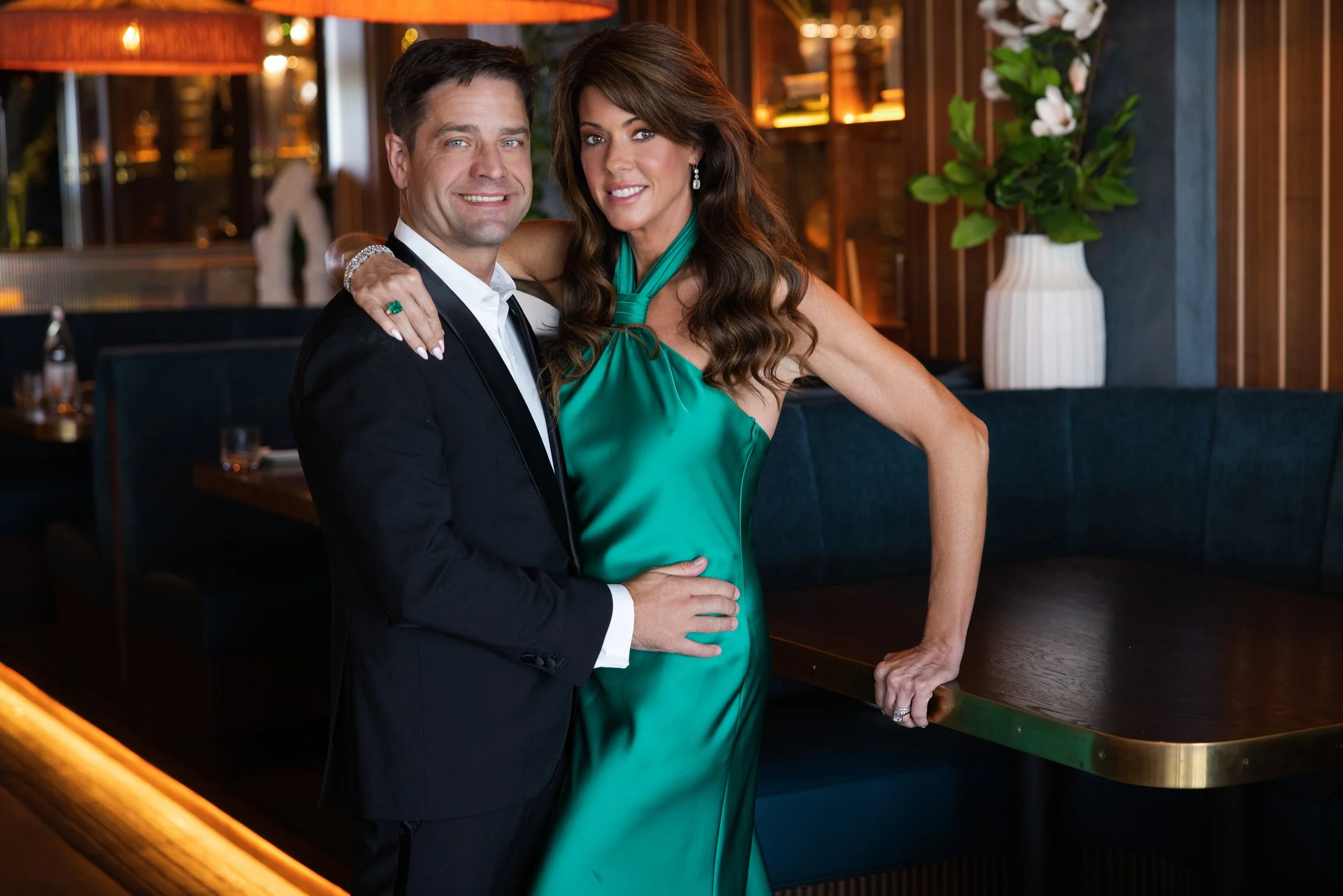A man and a woman in formal attire posing together in a warmly lit restaurant setting.