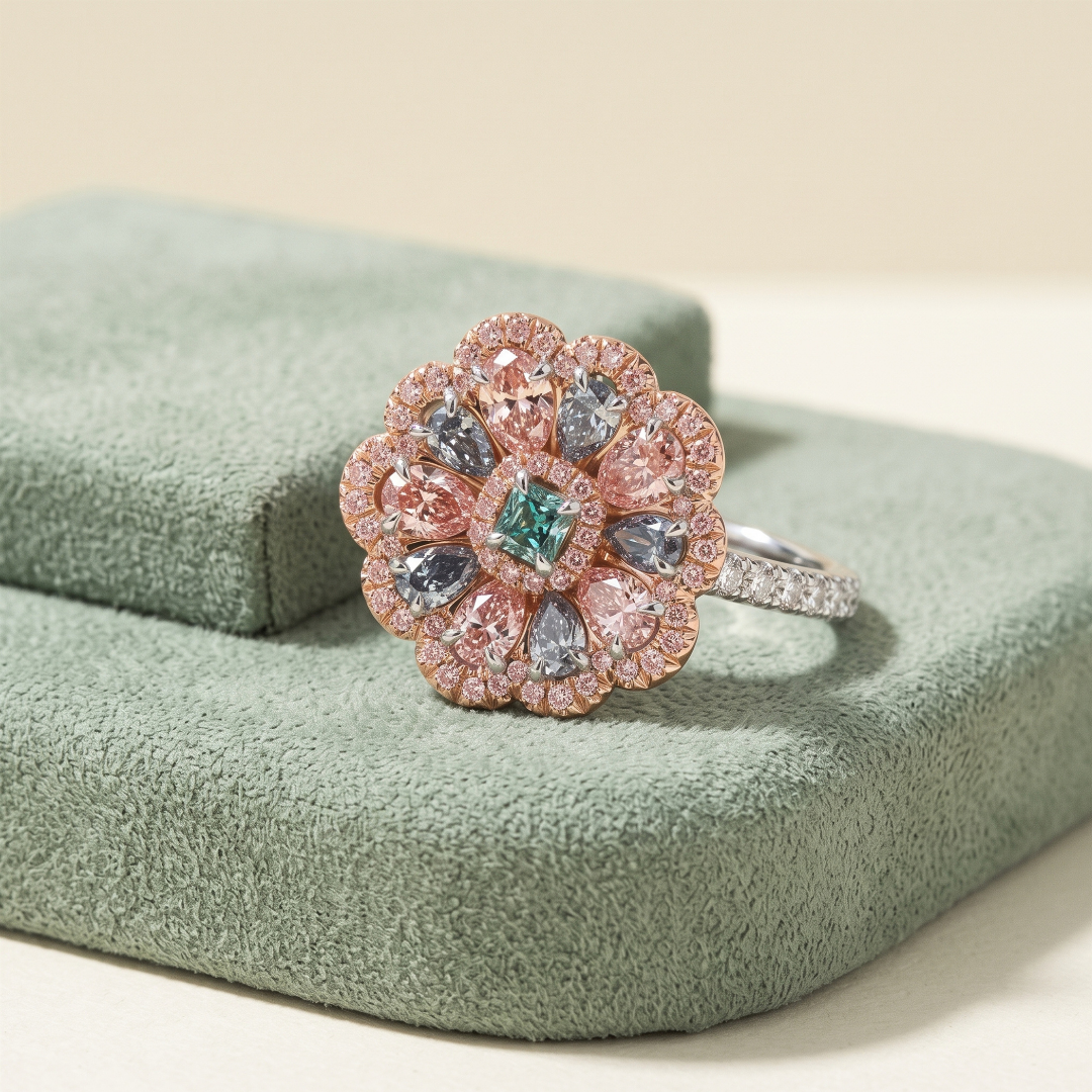 A floral-inspired ring with pink, gray, and green gemstones, set on a green textured jewelry box.