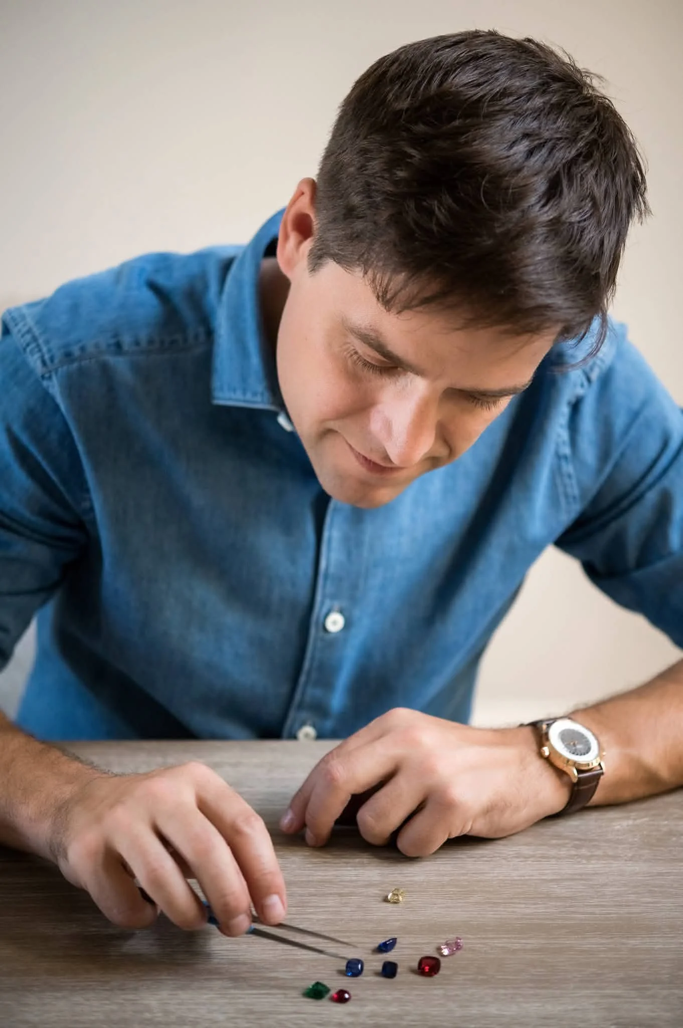 A man with dark hair wearing a blue shirt and a gold watch is sitting at a wooden table, arranging small colored gemstones and using tweezers.