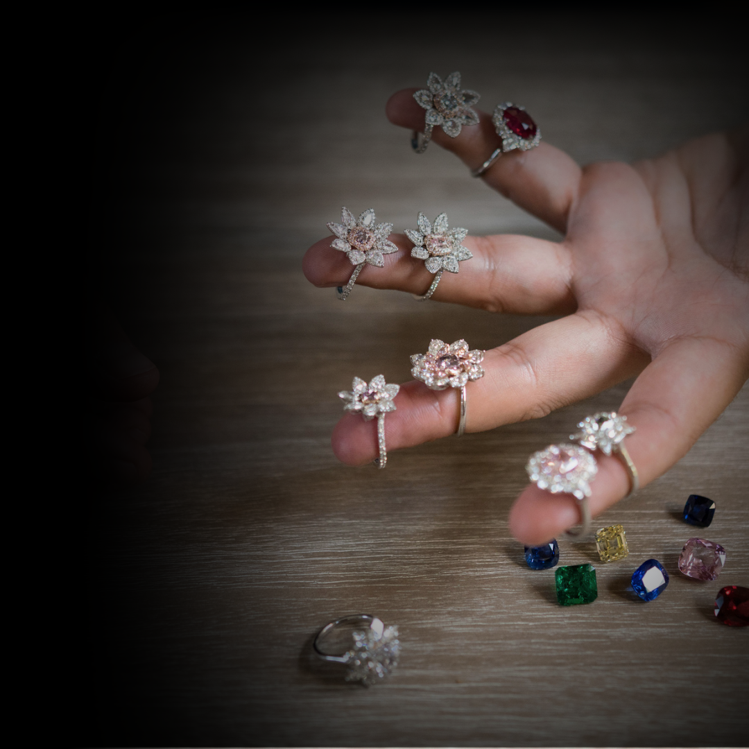 A hand displays various diamond and gemstone rings with floral and gemstone designs, surrounded by loose colorful gemstones on a wooden surface.