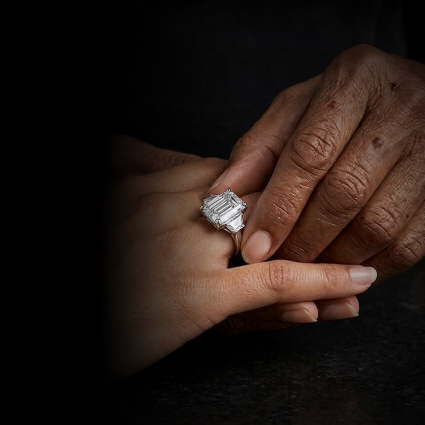Close-up of two hands, one with darker skin and one with lighter skin, gently touching. The hand with lighter skin is wearing a large, rectangular, diamond-studded ring.