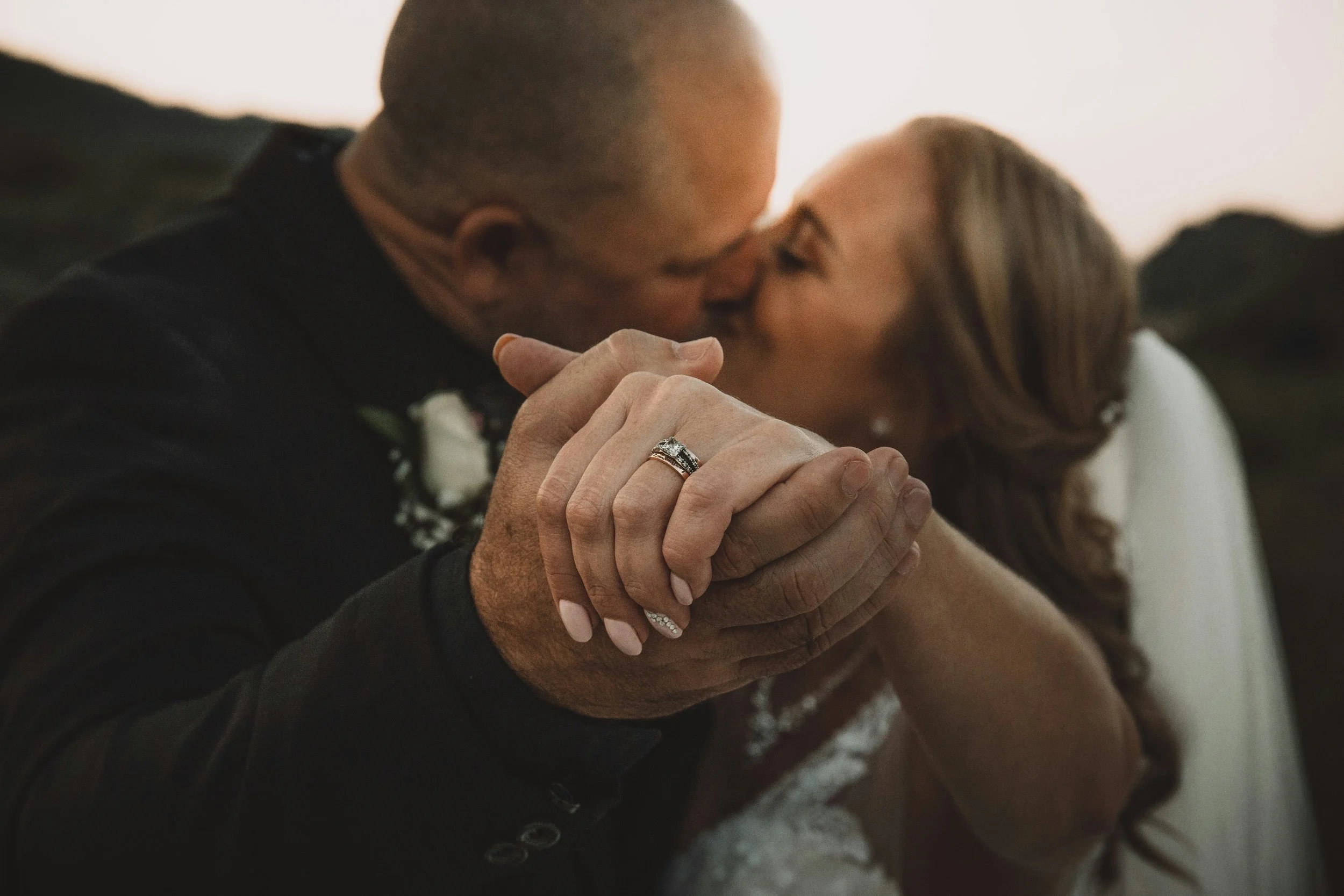 A close-up of a couple holding hands, with the man kissing the woman's forehead. The woman wears a wedding ring and a diamond bracelet; the man has a wedding band. The background is blurry, suggesting an outdoor setting during sunset.