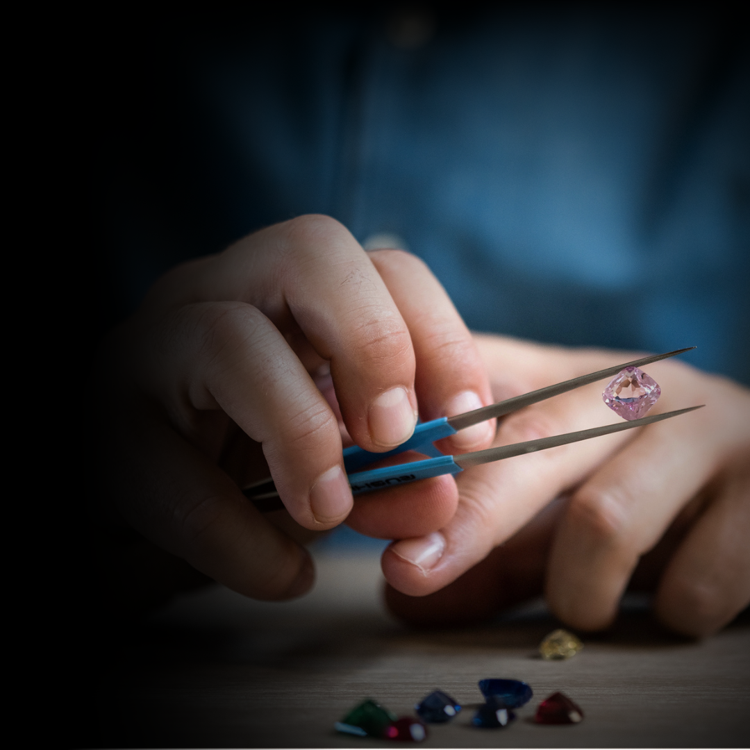 Person holding tweezers with a pink gemstone, surrounded by other colorful gemstones on a wooden surface.