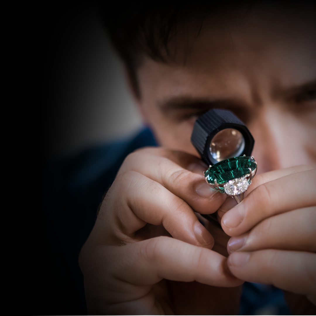 A person examining a ring with a large green gemstone and a diamond using a magnifying glass.