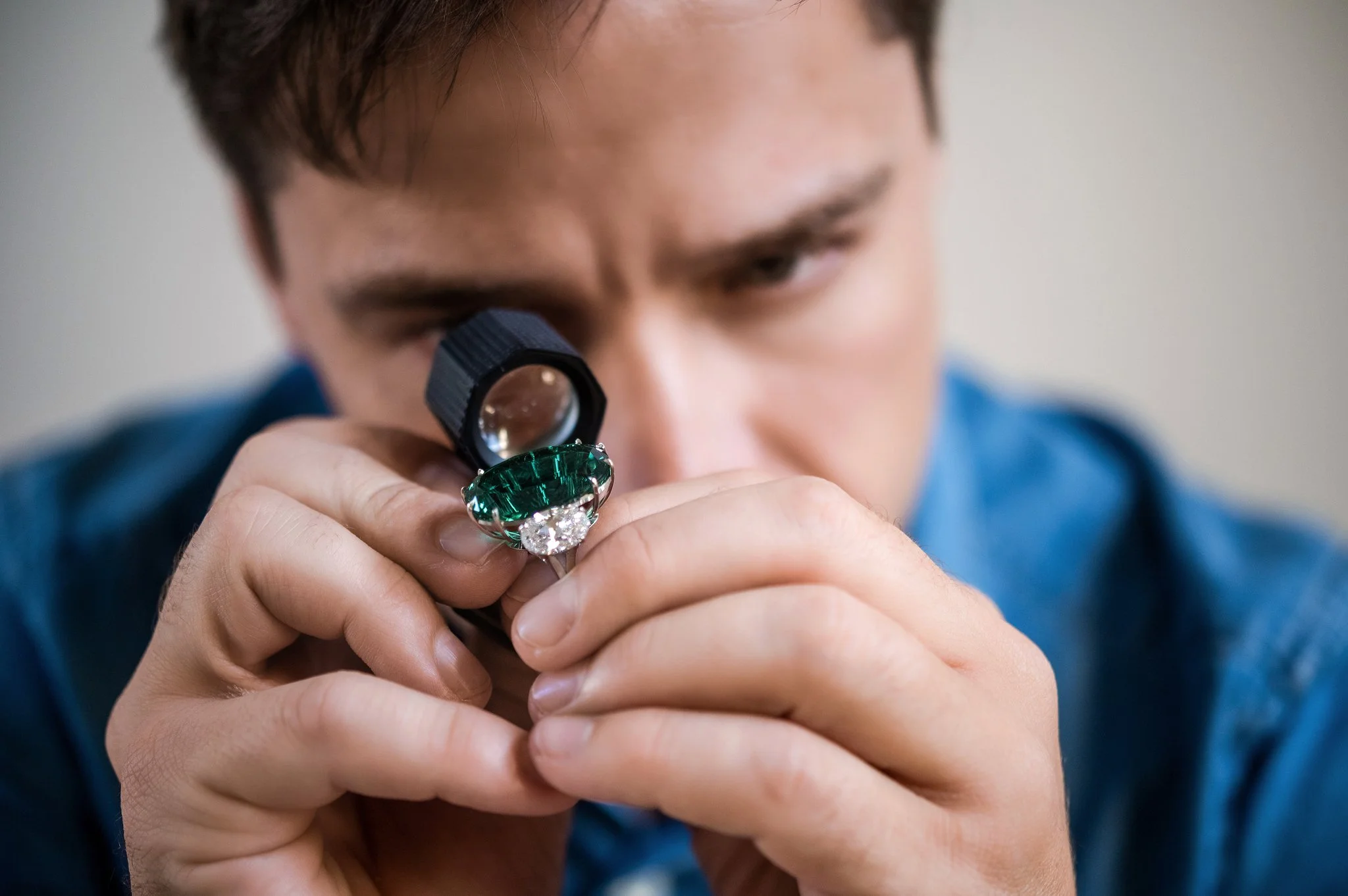 A person using a magnifying glass to closely examine a large green gemstone with a diamond in the center.