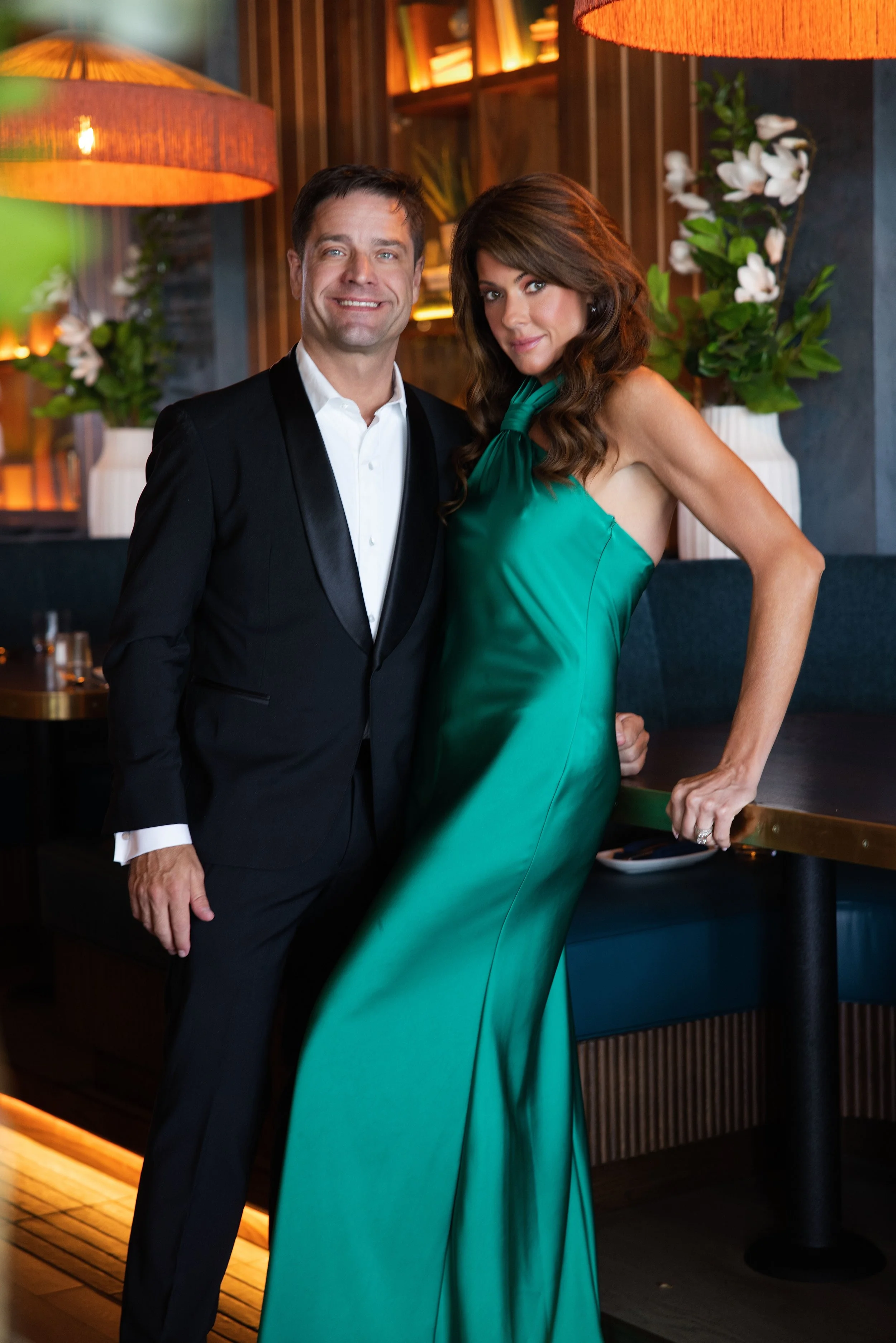 A man in a black tuxedo and a woman in a green satin gown pose together indoors, with warm lighting and floral decor in the background.