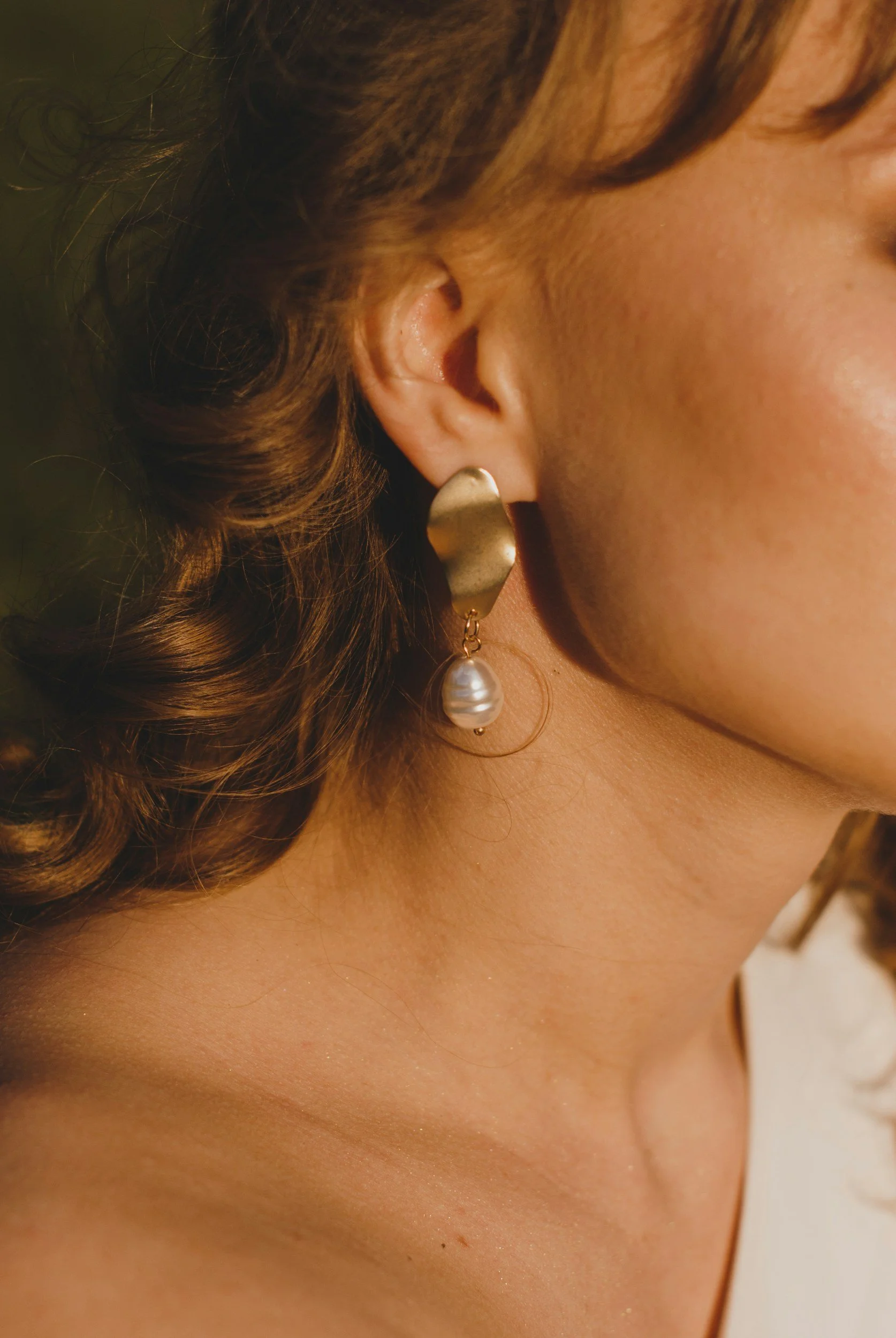 Close-up of a woman's face, showing her ear with a gold earring featuring a large pearl, with curly brown hair and smooth skin.
