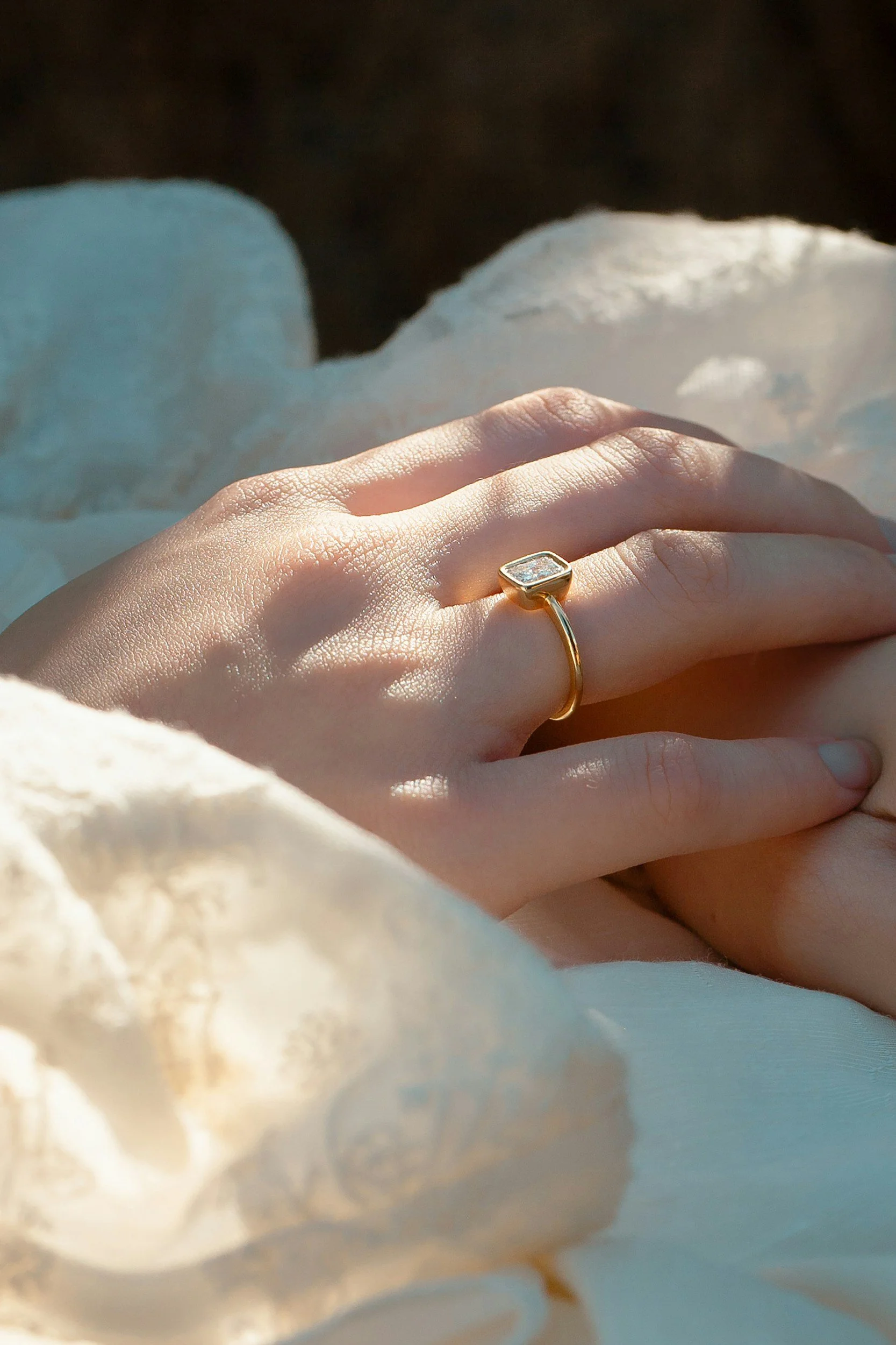 Close-up of a hand wearing a gold ring with a rectangular gemstone, resting on a soft, light-colored fabric.