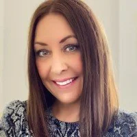 A woman with long brown hair, smiling, wearing a patterned sweater, indoors with a neutral background.