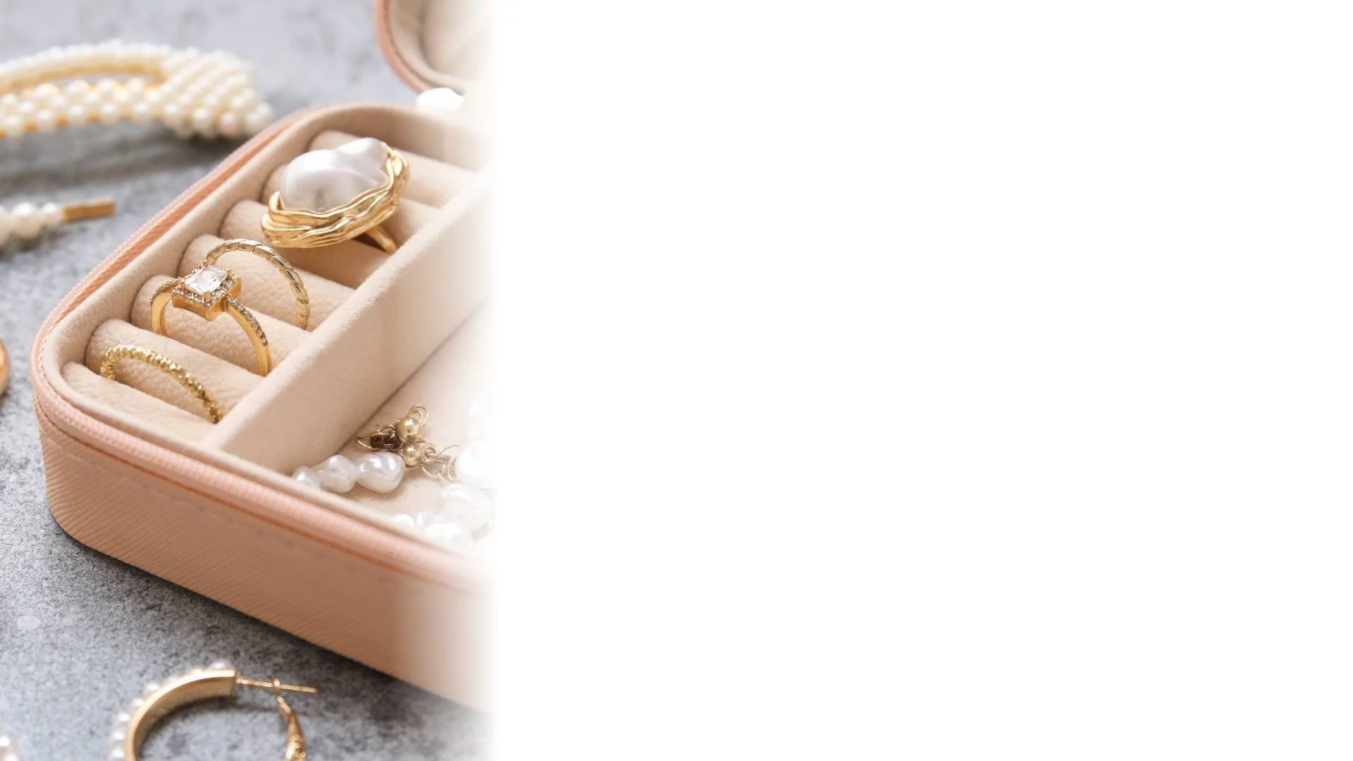 Close-up of a beige jewelry box containing gold rings and a pearl ring, with pearl earrings and additional jewelry scattered around on a gray surface.