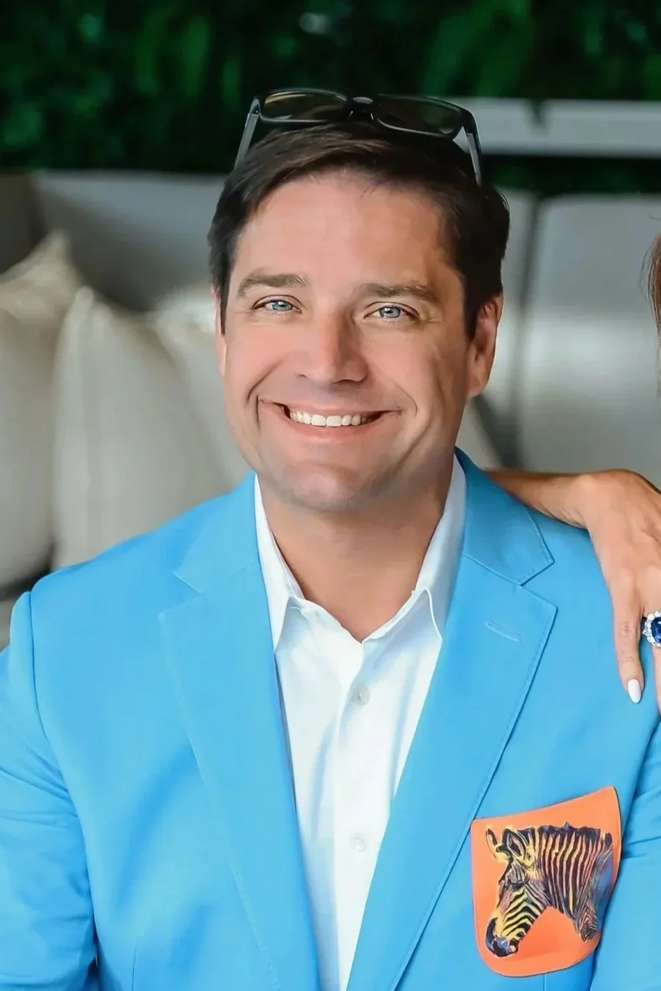 Man with tan skin, dark brown hair, and blue eyes smiling, wearing a bright blue blazer with a patch of a zebra on the pocket, white shirt, and sunglasses on his head.
